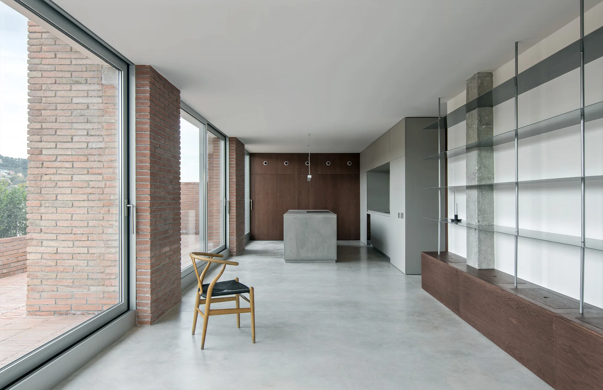 A minimalist kitchen with a grey concrete floor and island, tall sliding glass doors framed by brick columns, and a wood shelving unit.