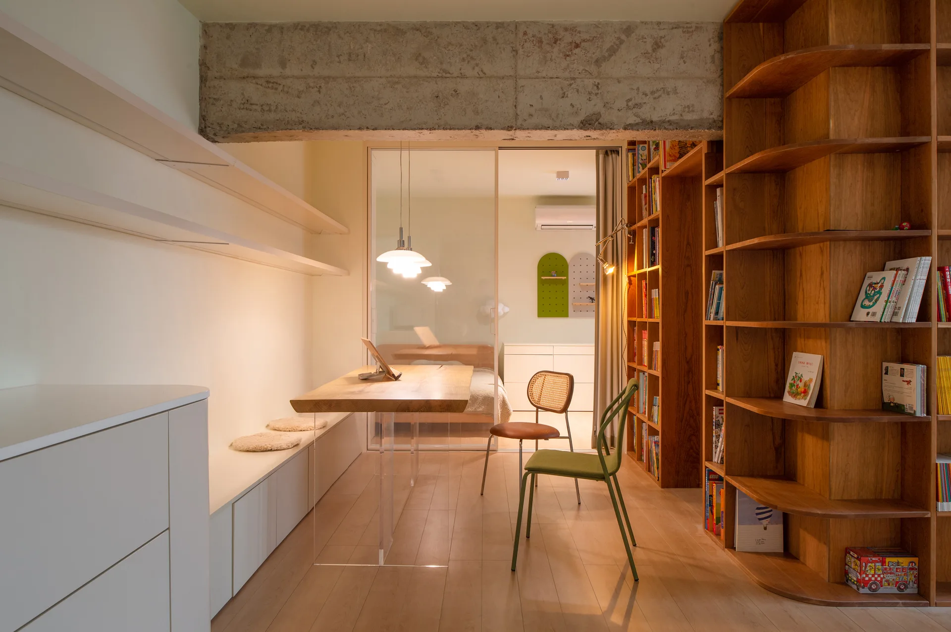 A modern home office featuring a thick wood desk with clear acrylic legs, built-in wooden bookshelves, and an exposed concrete ceiling beam.