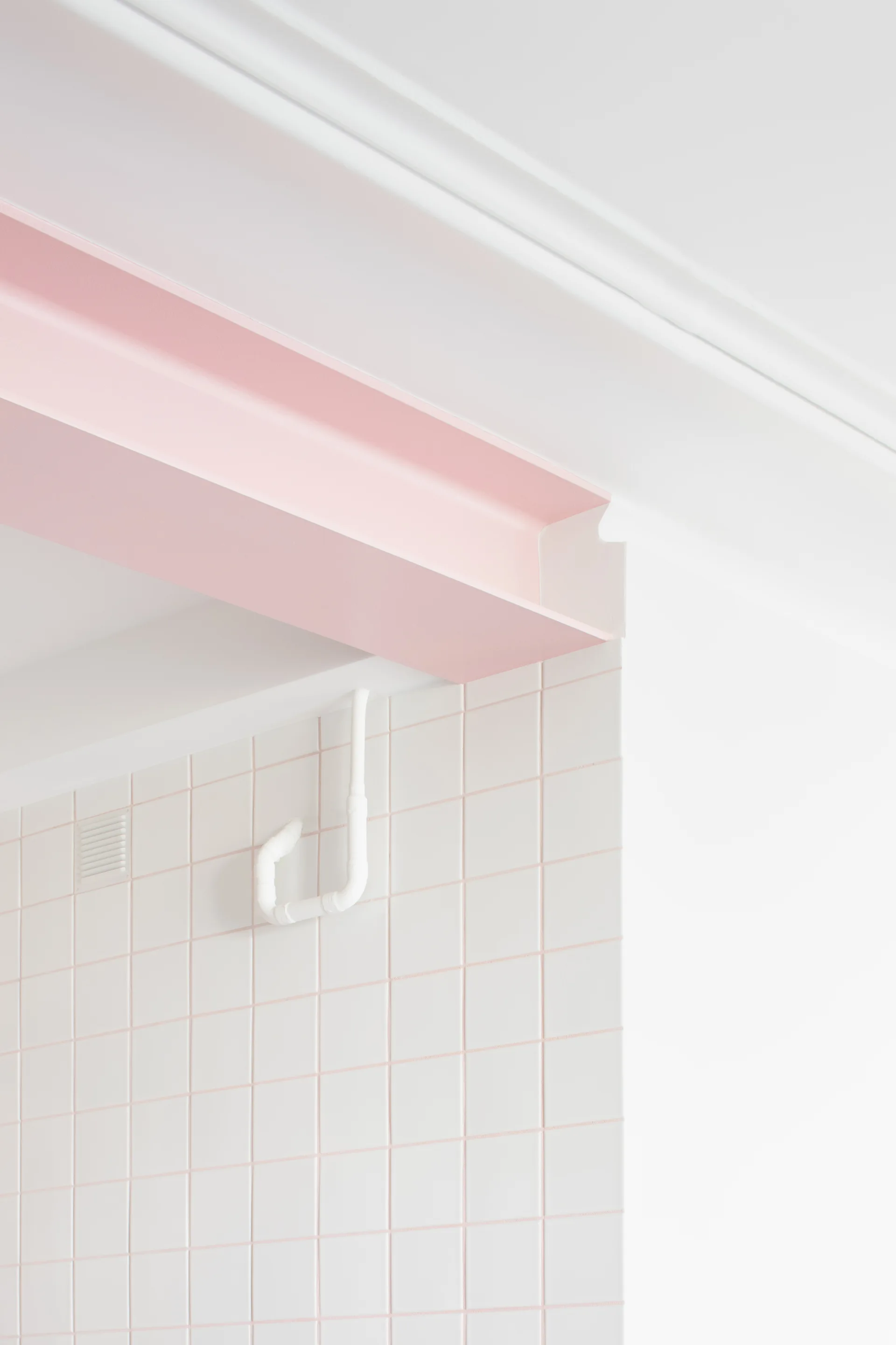 A pale pink painted structural steel beam meets a white square grid tiled wall and white ceiling.