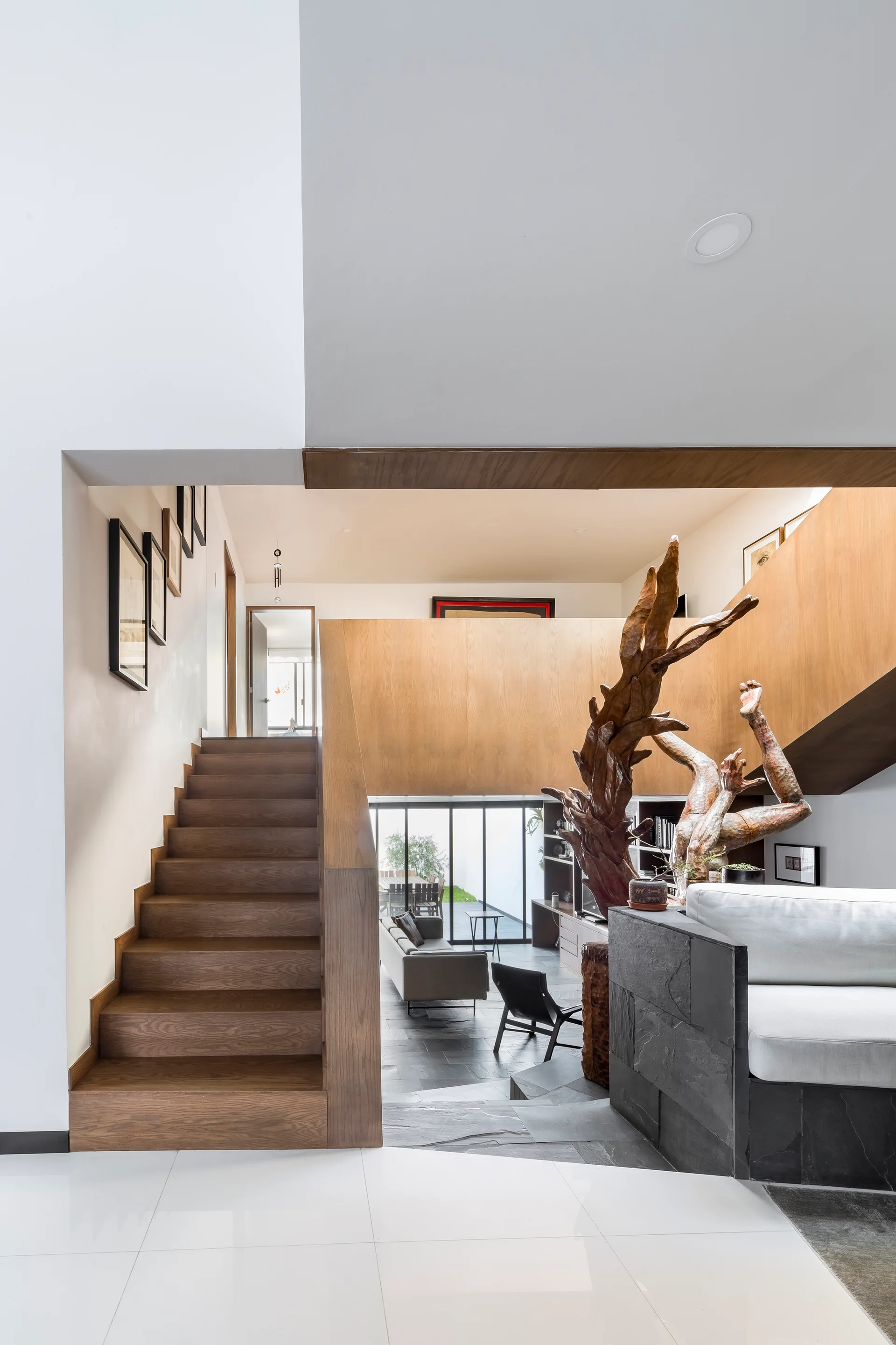 A wooden staircase in a split-level home leads down to a living area with dark slate tile flooring, a grey sofa, and a large tree-root sculpture.