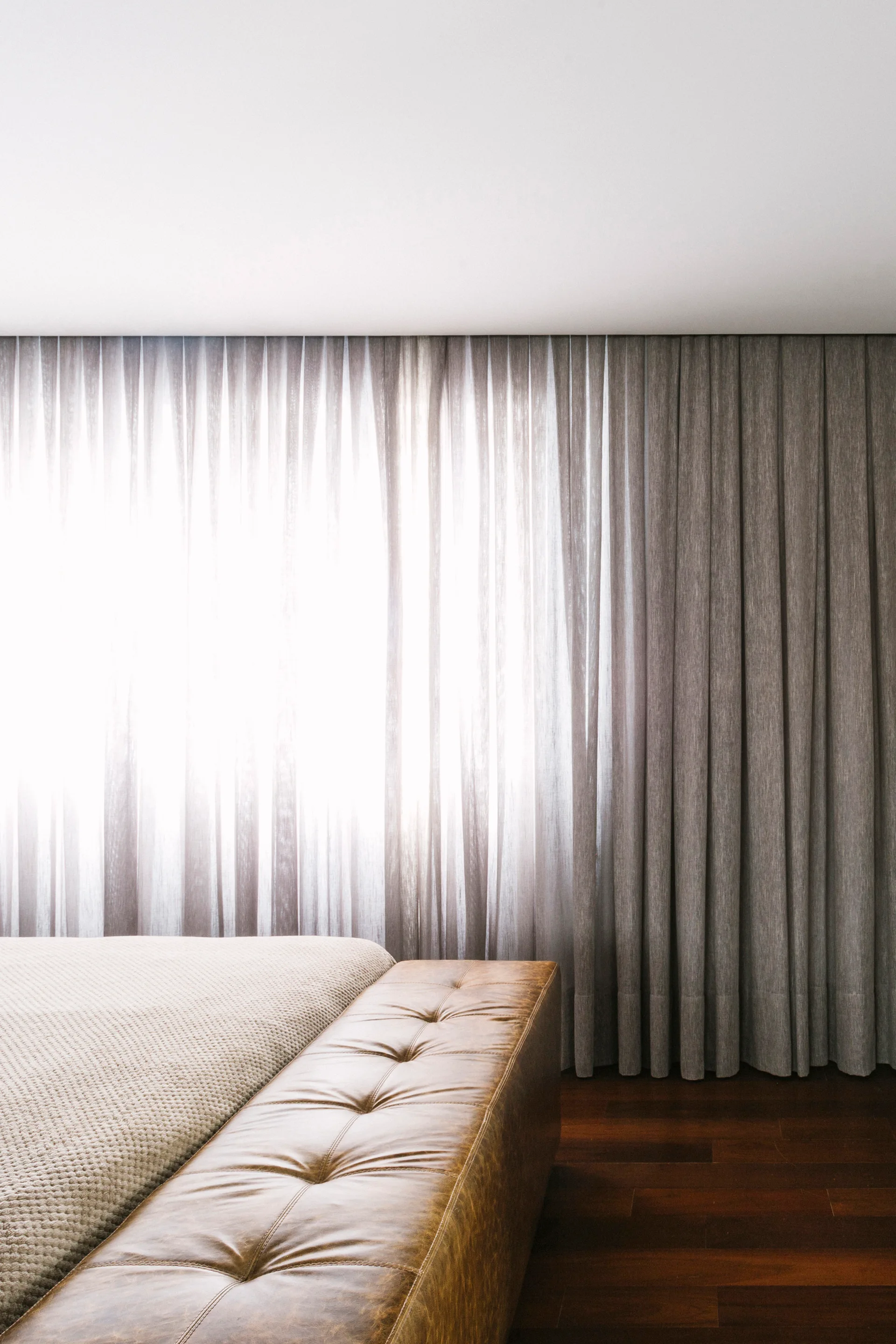A tufted brown leather bench stands at the foot of a bed in front of floor-to-ceiling grey curtains and dark wood floors.