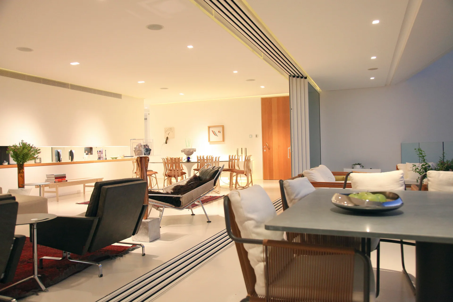 An open-plan living and dining room features white floors, recessed ceiling lights, mid-century style chairs, and metal tracks for a sliding partition wall.