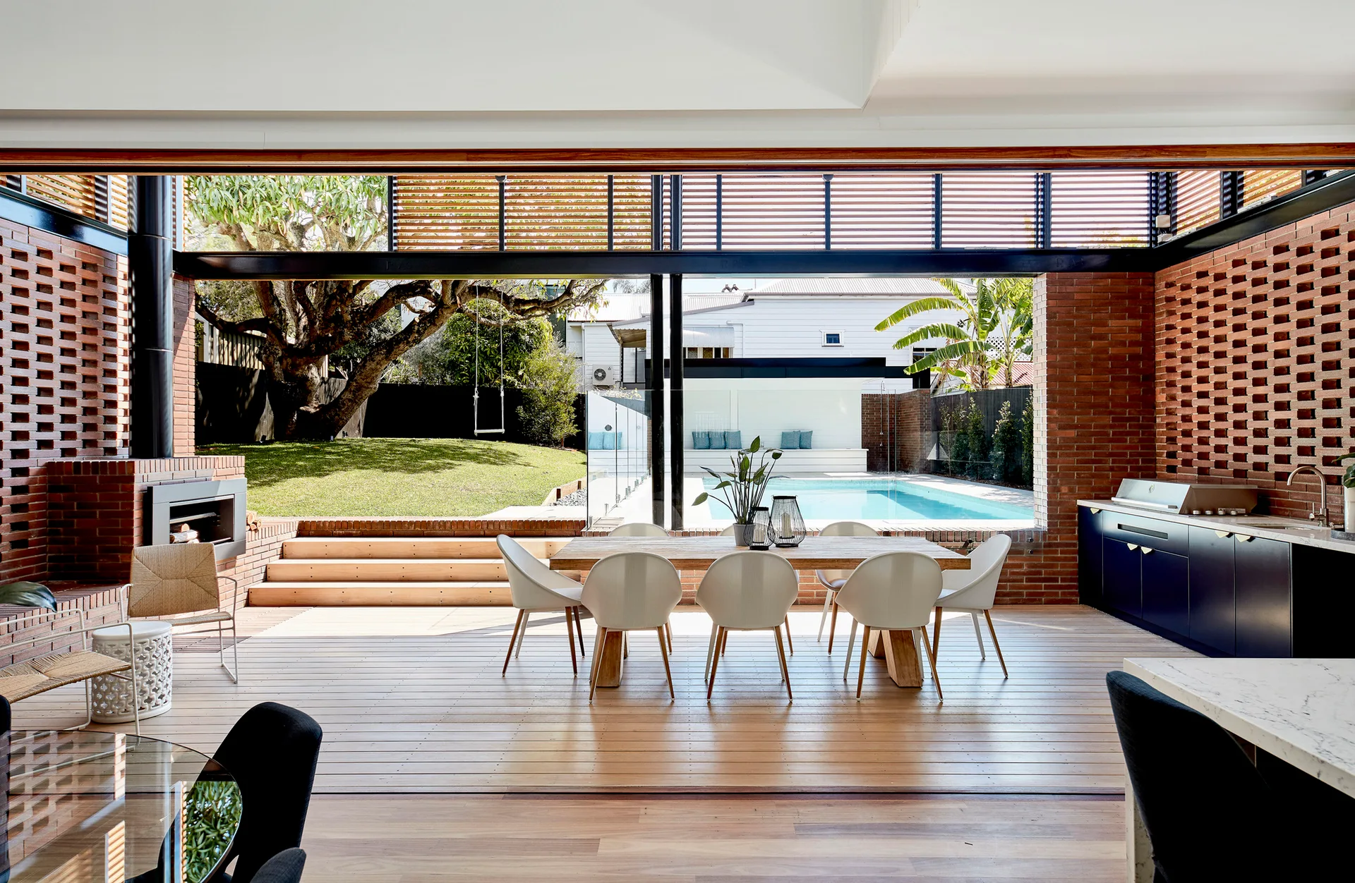 An open-air covered deck features a wooden dining table, white chairs, navy outdoor kitchen cabinets, and brick masonry walls overlooking a pool.