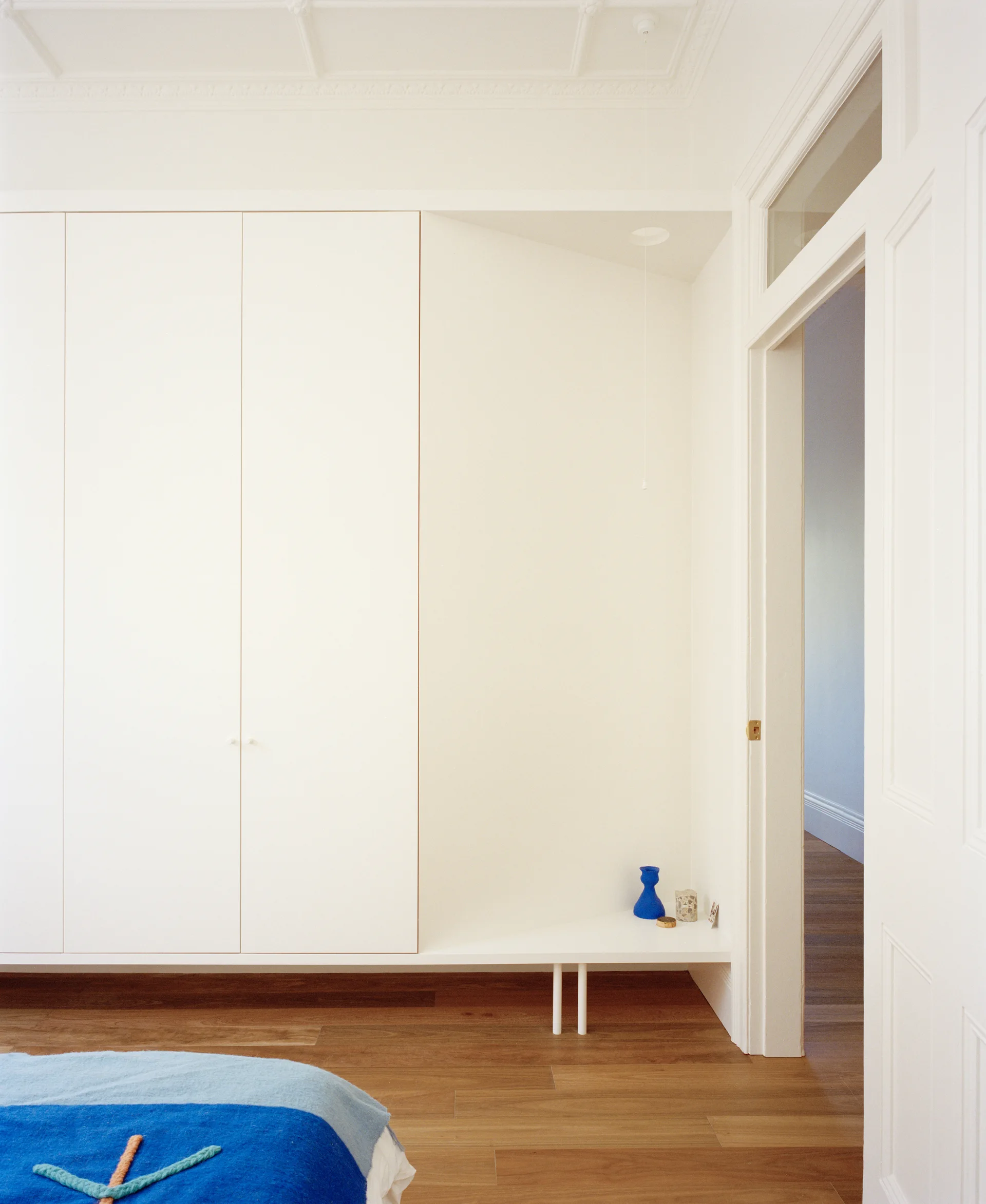 A minimalist white built-in wardrobe stands next to an open niche with a small blue vase, set against hardwood floors and an ornate ceiling.