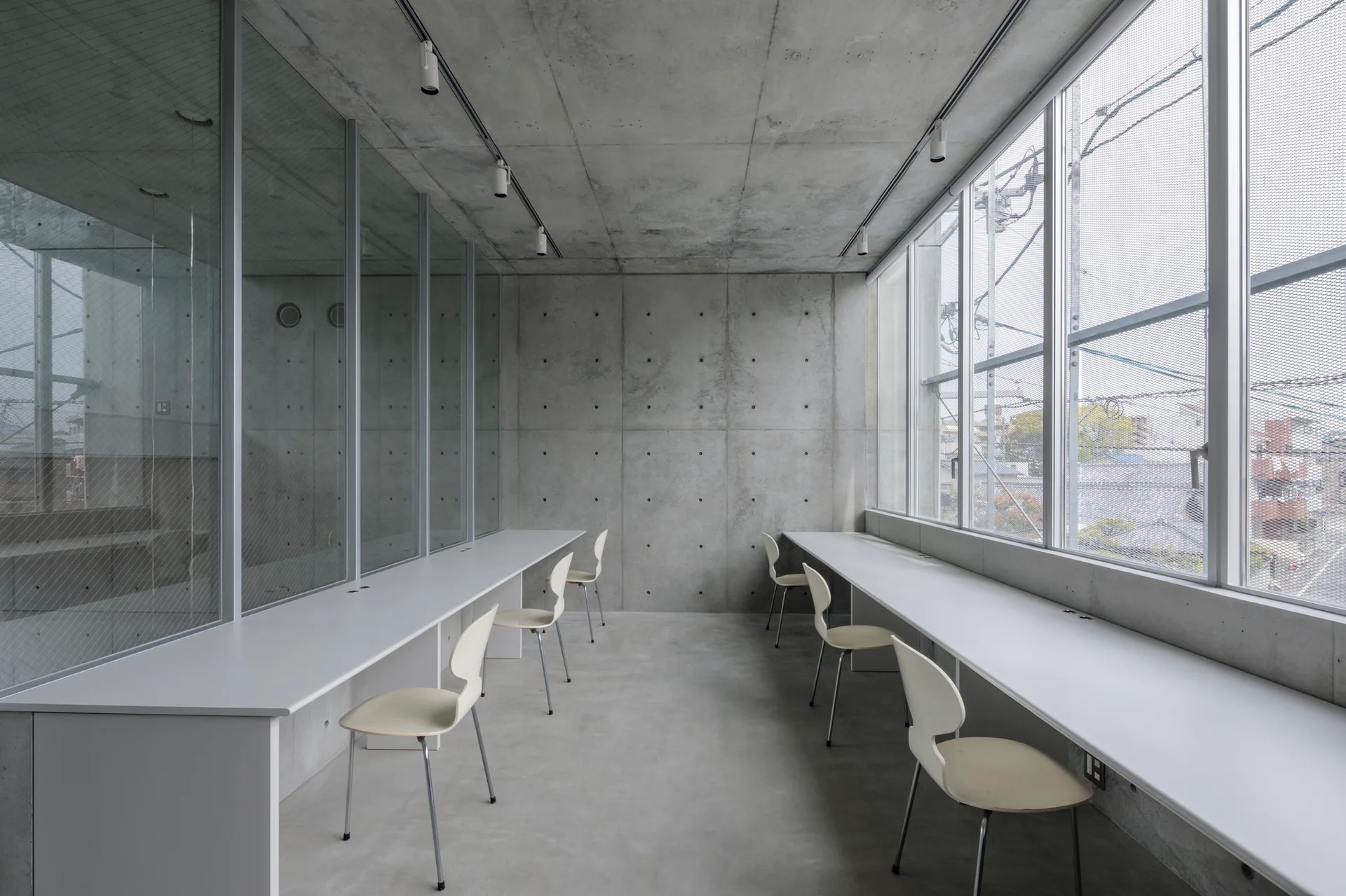 A minimalist industrial workspace features long white desks and cream chairs set against raw exposed concrete walls, a concrete floor, and glass partitions.