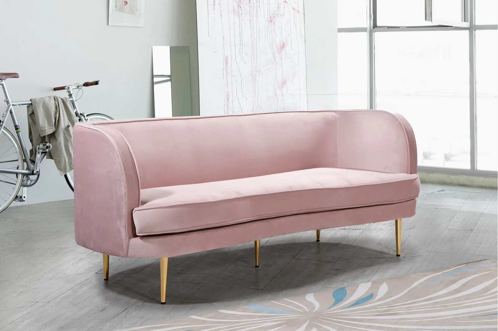 A pink velvet curved sofa with gold legs stands on a light grey wood floor next to a bicycle and an abstract painting.