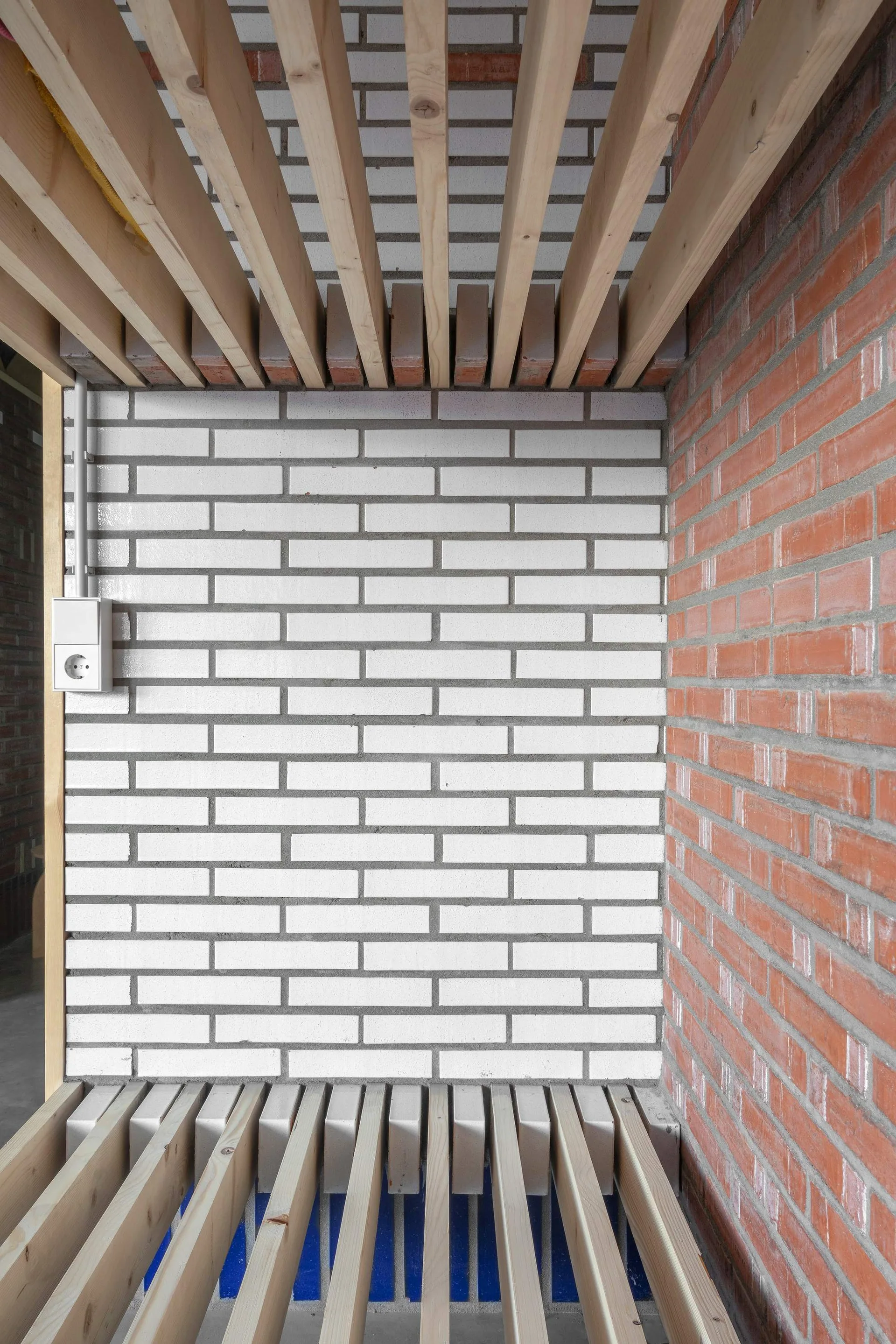 An industrial interior corner showing a white painted brick wall next to a red clay brick wall with parallel light wood slats at the top and bottom.