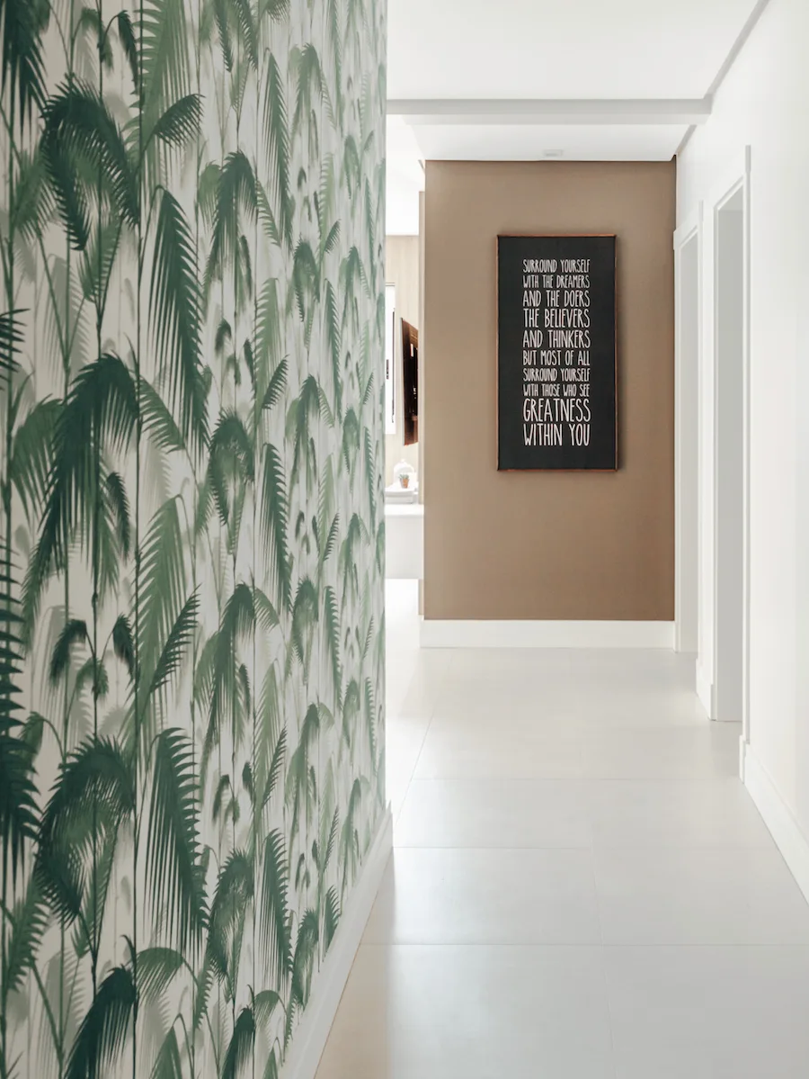 A bright hallway featuring green palm leaf wallpaper on the left wall and a framed motivational quote on a taupe accent wall at the end of the corridor.