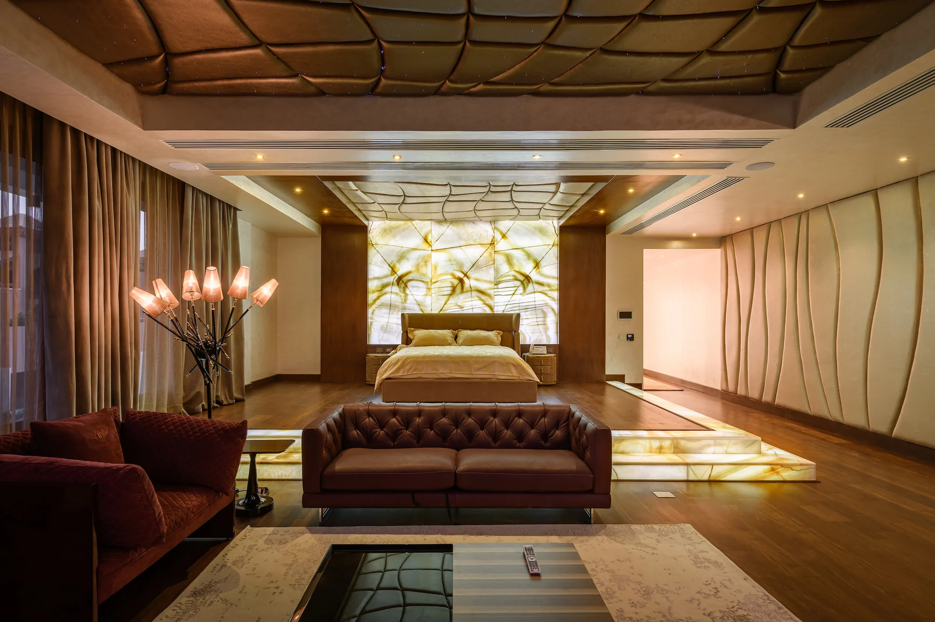 A bedroom features a raised wooden platform for the bed, a backlit glowing onyx feature wall, and a tufted leather sofa in the foreground.
