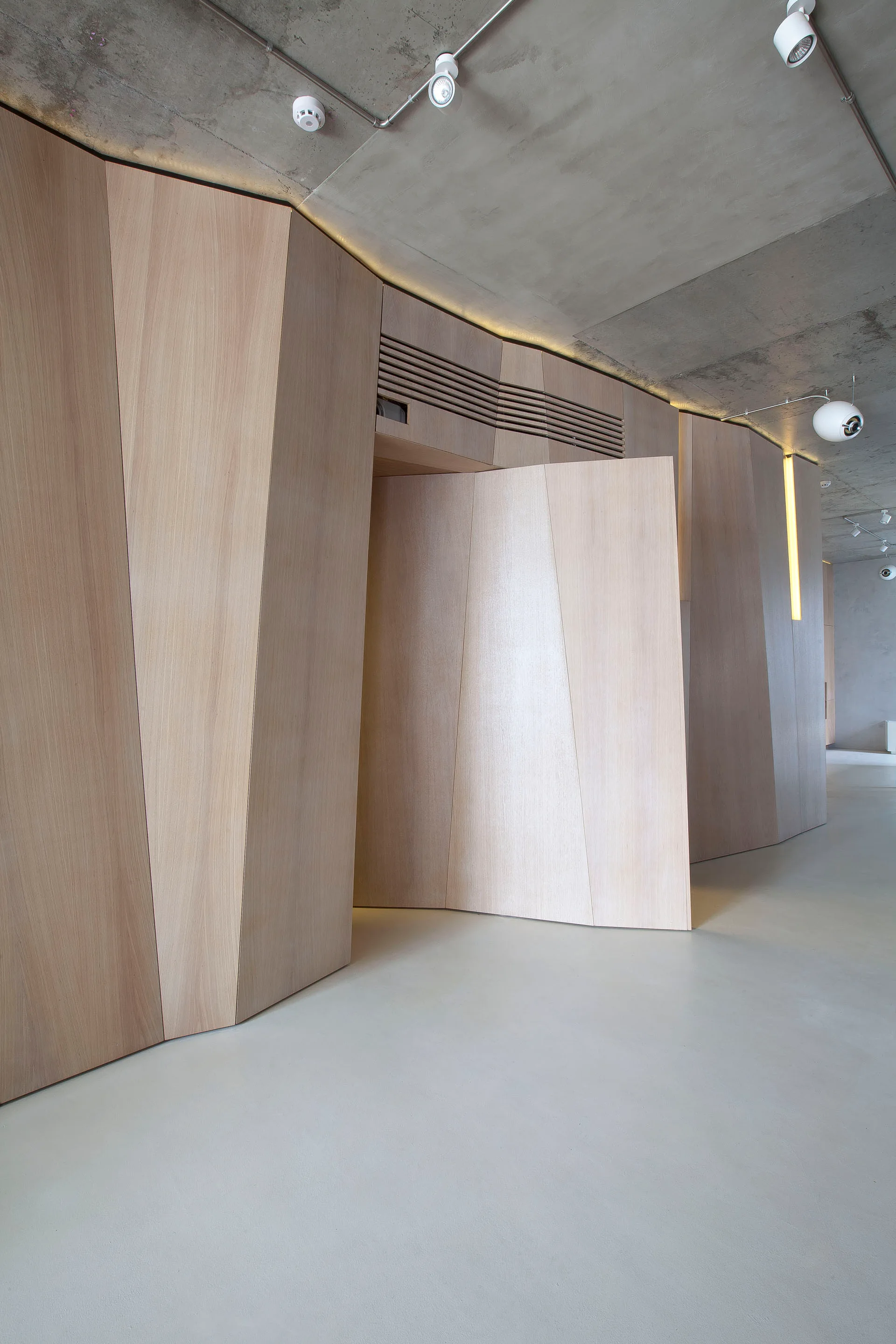A minimalist interior featuring geometric light wood wall panels with a pivoting door under a raw concrete ceiling.