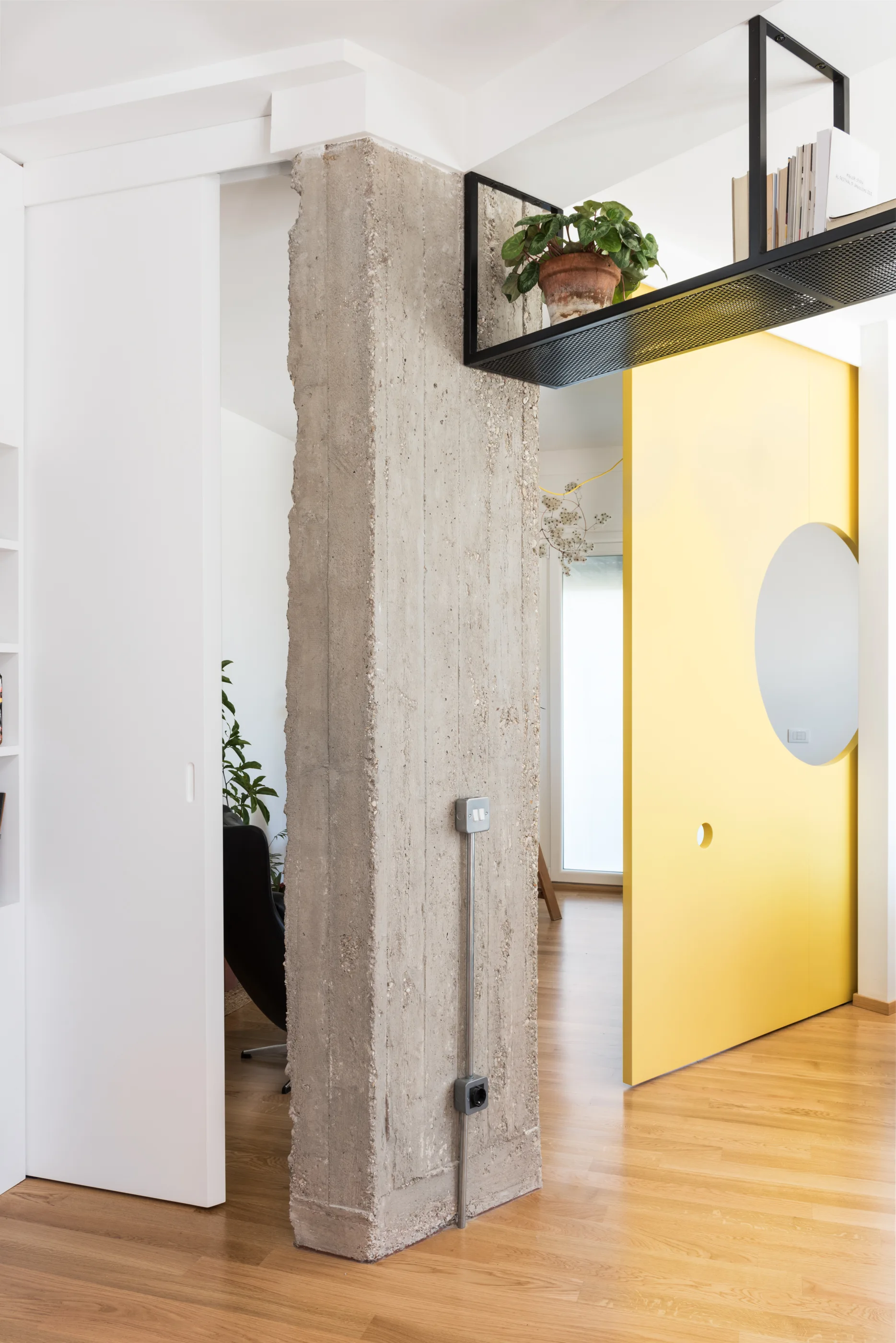 An interior view shows an exposed raw concrete column, a sliding bright yellow door with a circular cutout, and a suspended black metal shelf.