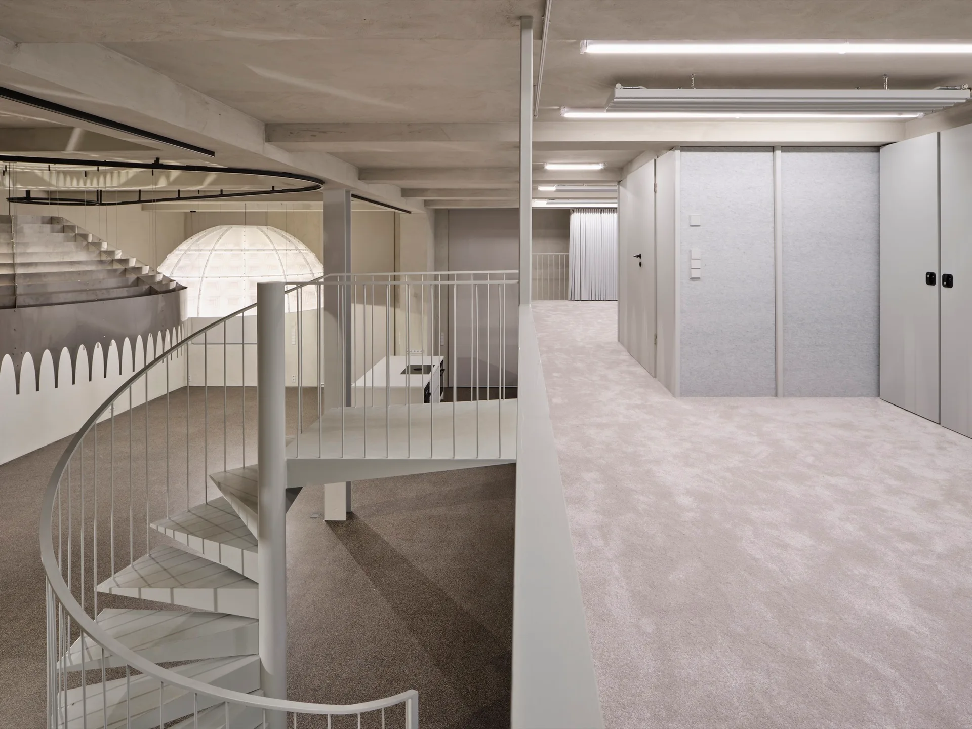 A white metal spiral staircase leads to a grey carpeted mezzanine landing beneath an exposed concrete ceiling with minimalist cabinets.