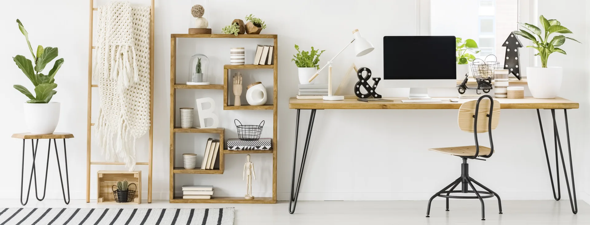 A bright home office features a wooden desk with black hairpin legs, a matching open shelving unit, and a wooden swivel chair against a white wall.