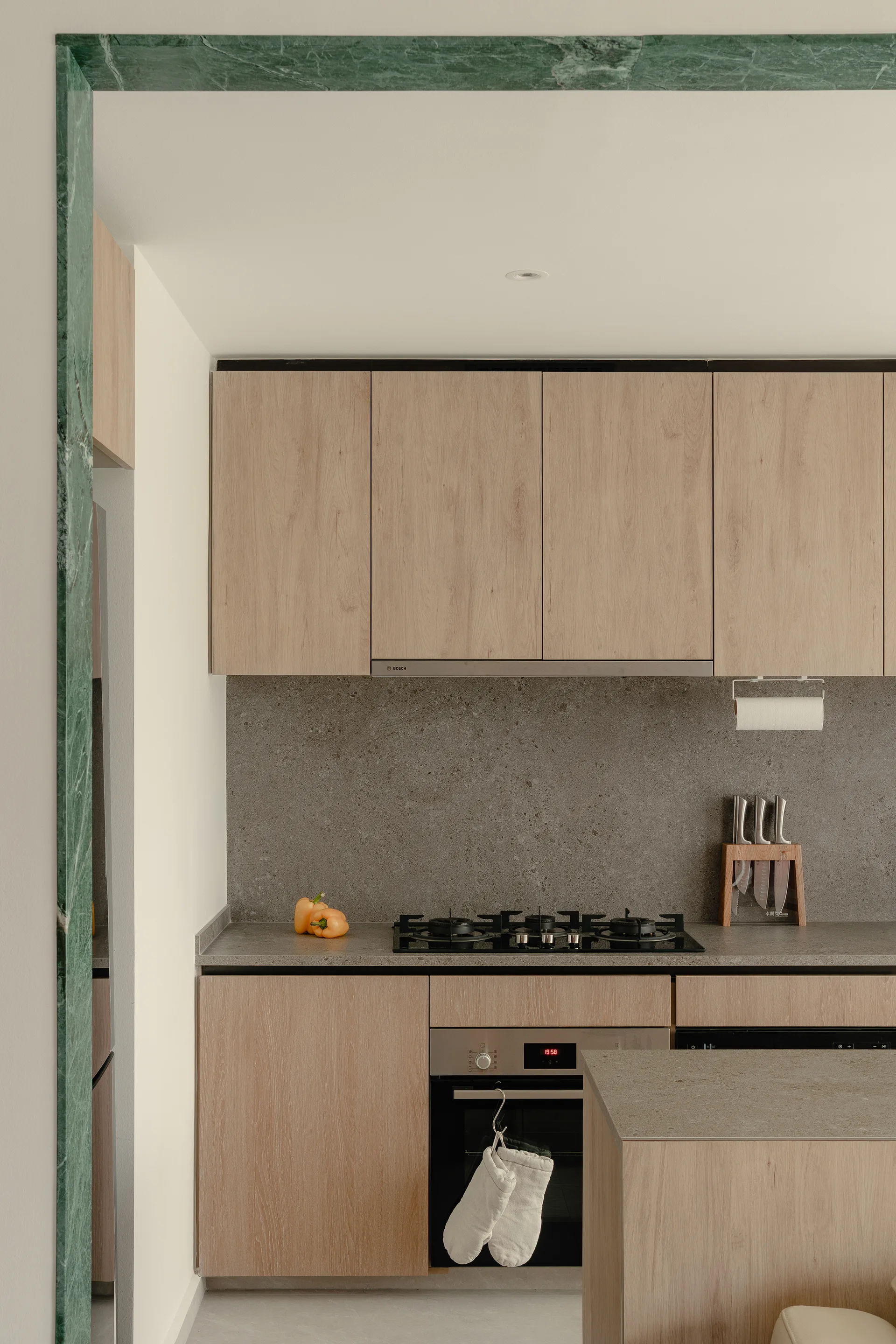 A minimalist kitchen featuring light wood cabinets, grey stone countertops, and a built-in oven, framed by a green marble entryway arch.