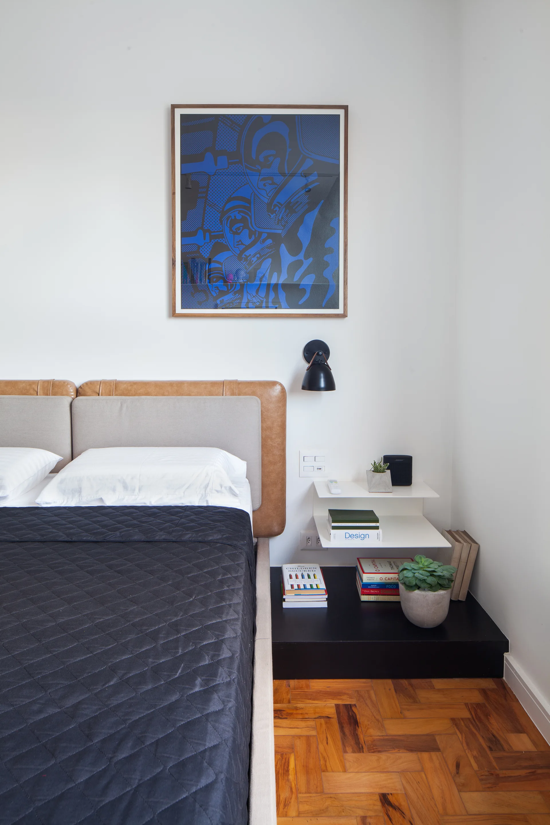 A modern bedroom corner featuring a bed with a leather and fabric headboard next to a tiered white nightstand on herringbone wood flooring.