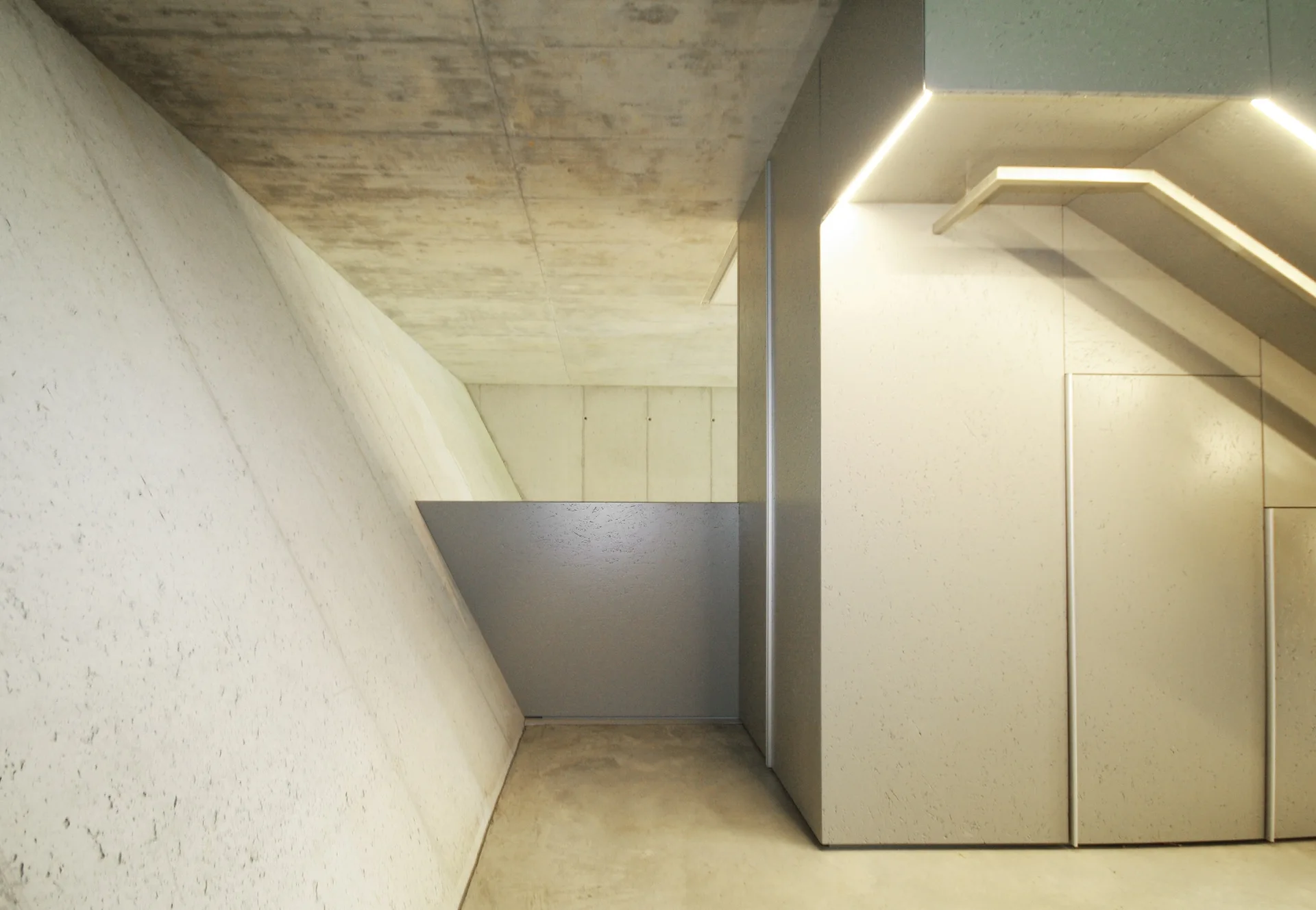 A minimalist hallway featuring raw concrete walls, a smooth floor, and an integrated staircase with flush cabinets and built-in LED lighting.