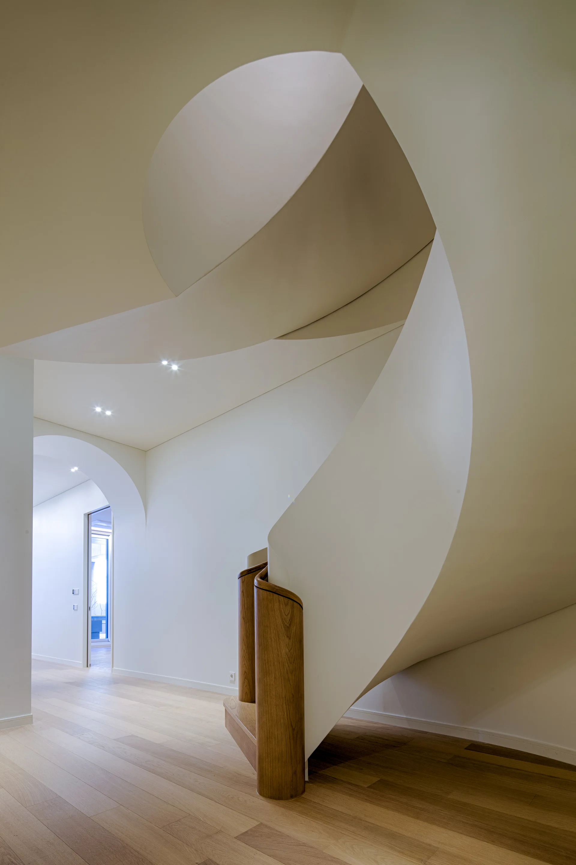 A sculptural white spiral staircase with a curved wooden handrail stands on light wood floors near a white plaster archway.