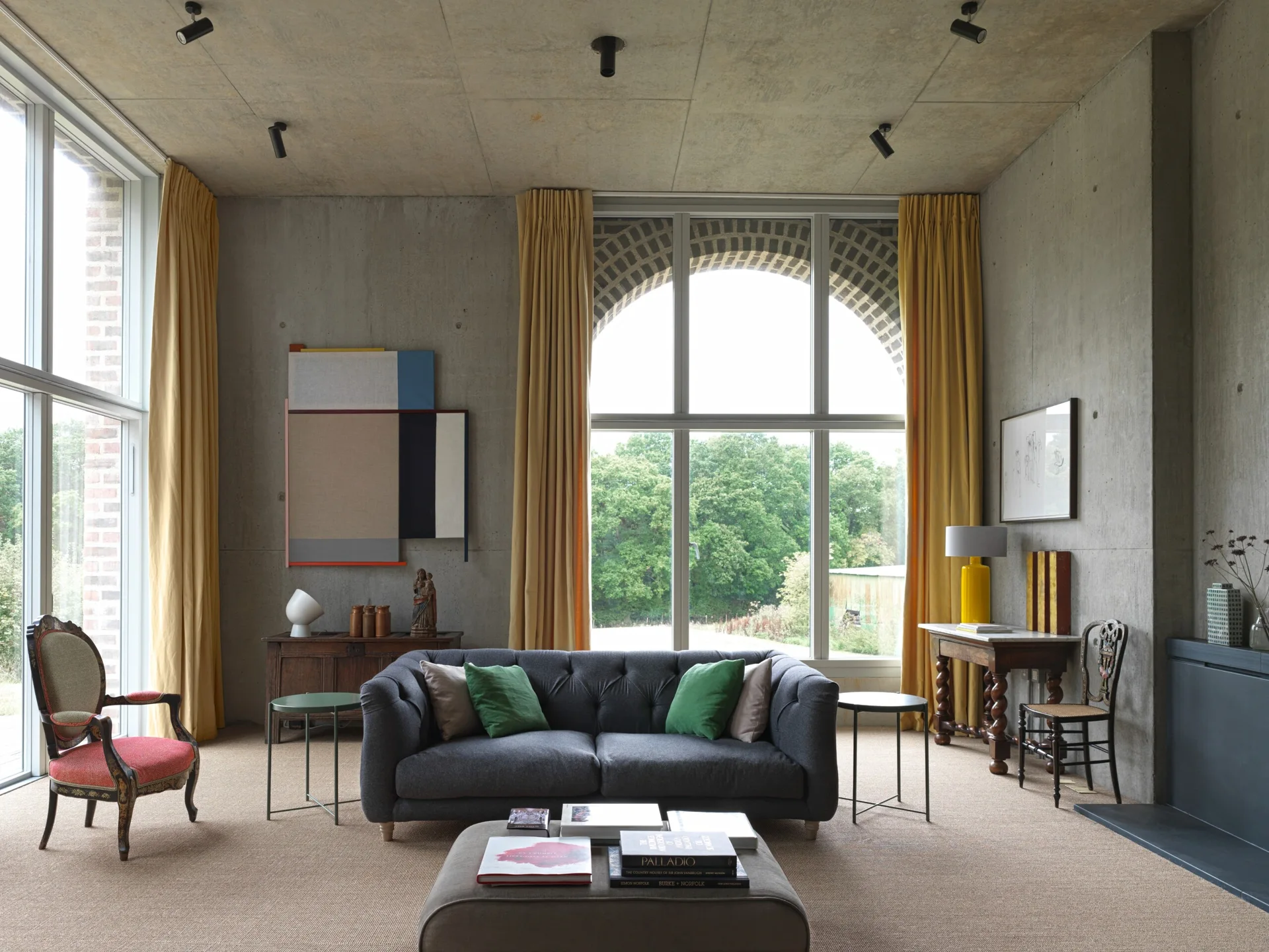 An eclectic living room featuring raw concrete walls, a grey tufted sofa with green pillows, yellow drapery, and a large arched window.