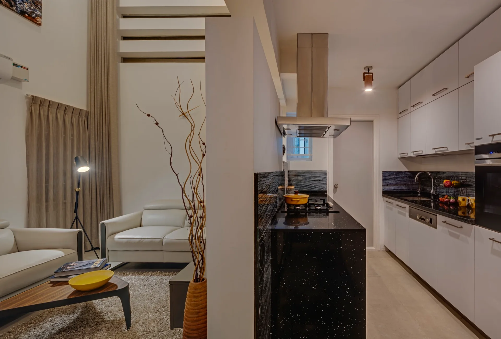 A modern kitchen with black stone countertops and white cabinets sits adjacent to a cozy living area with a white leather sofa and shag rug.