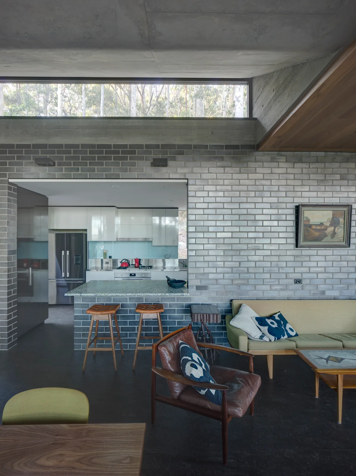 An open-plan living space features a grey brick wall, concrete ceiling, a low-profile sofa, and a kitchen visible through a rectangular cutout.