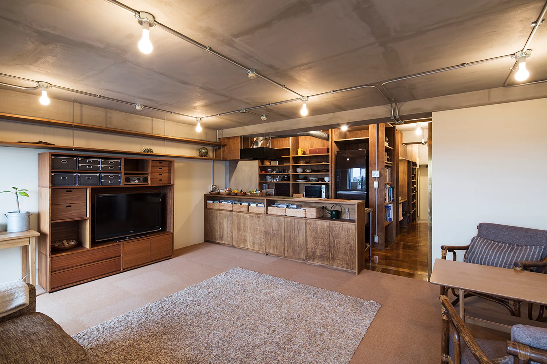 An open concept living room features warm wood cabinetry, a tan shag rug, and an exposed raw concrete ceiling with metal conduit wiring.