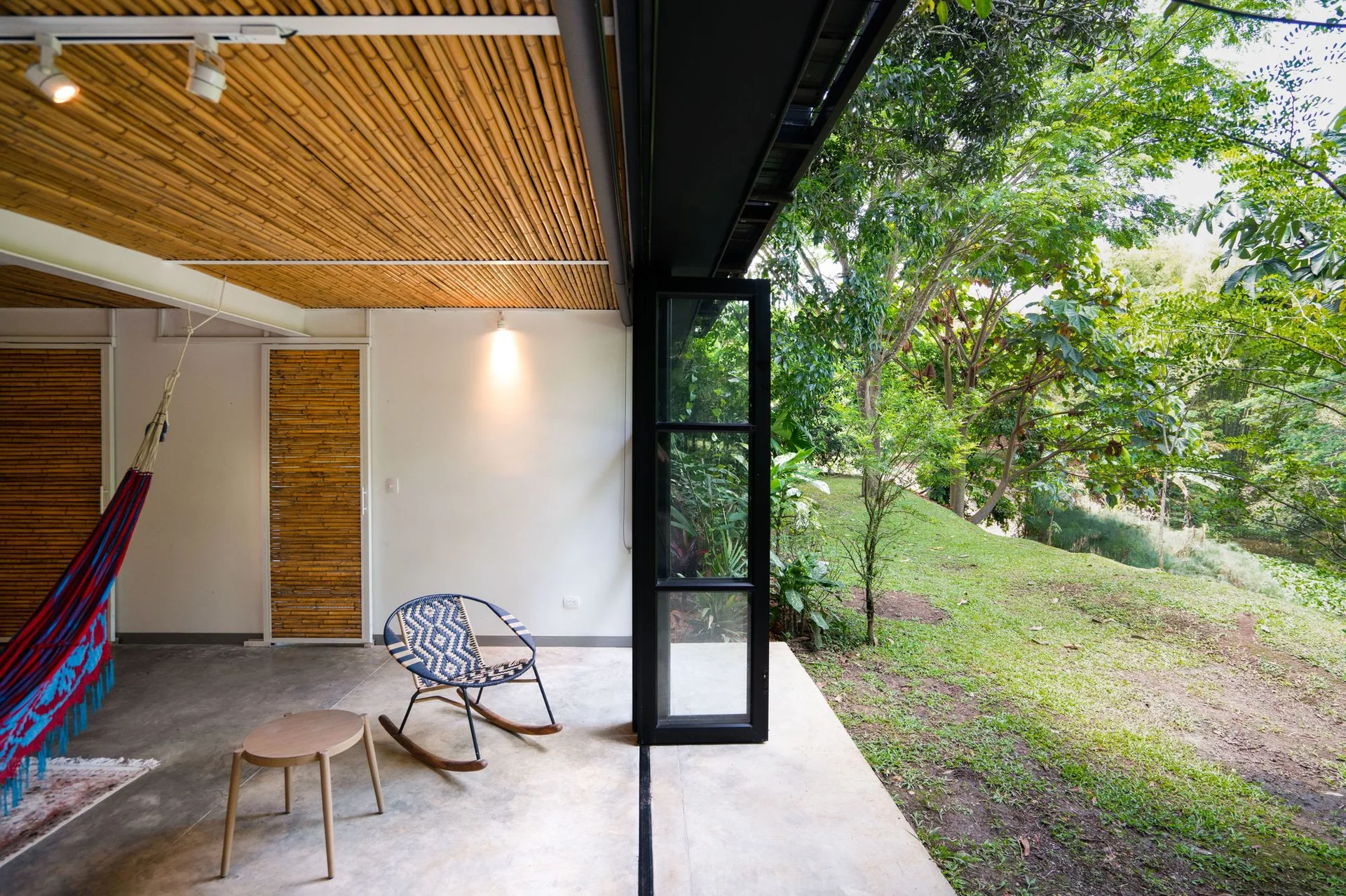 A concrete patio featuring a slatted bamboo ceiling, a woven rocking chair, and a black glass folding door that opens to a green lawn.