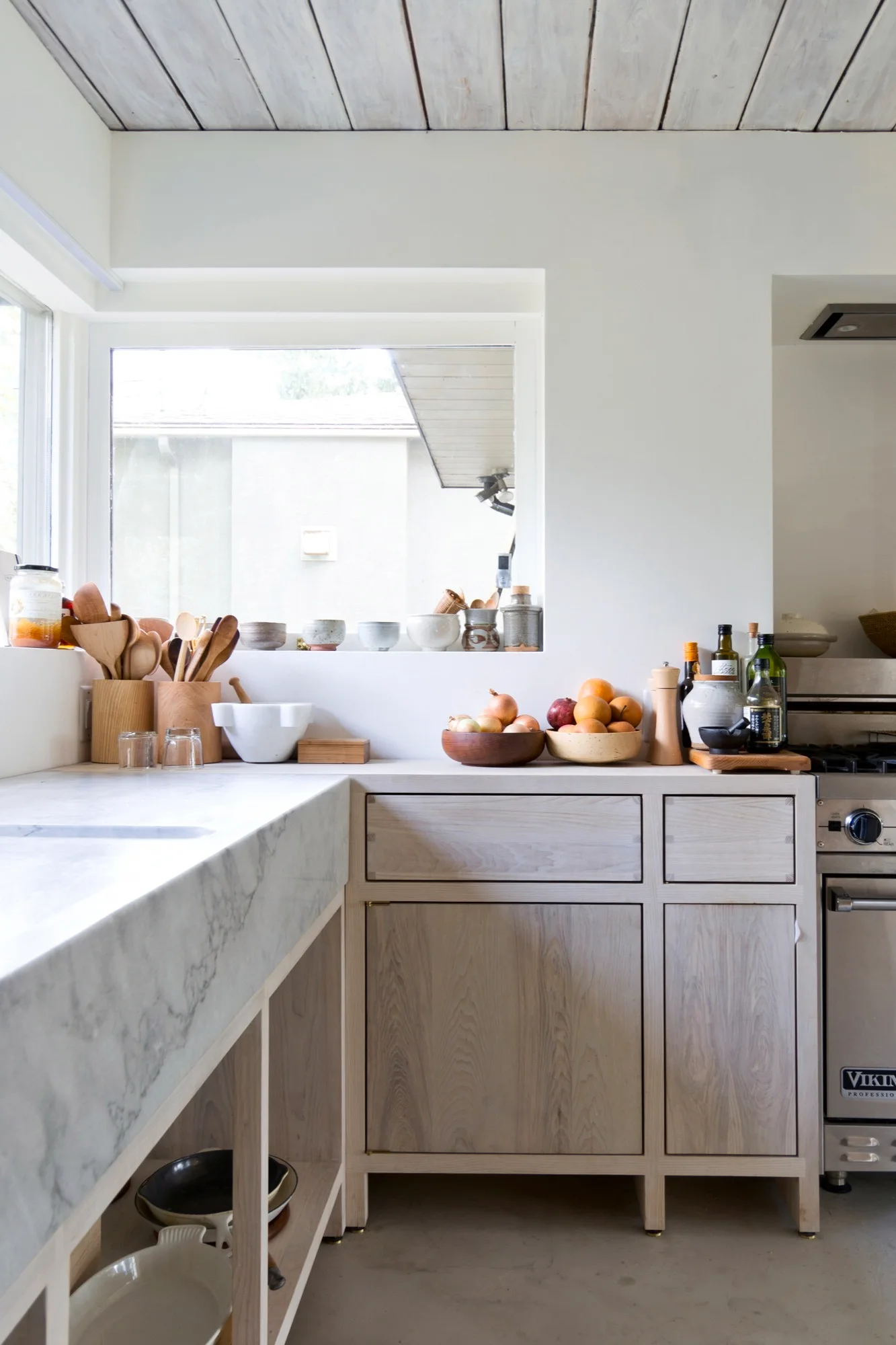 A bright kitchen features pale wooden base cabinets, a thick marble countertop, a wood-plank ceiling, and a deep window sill with small bowls.