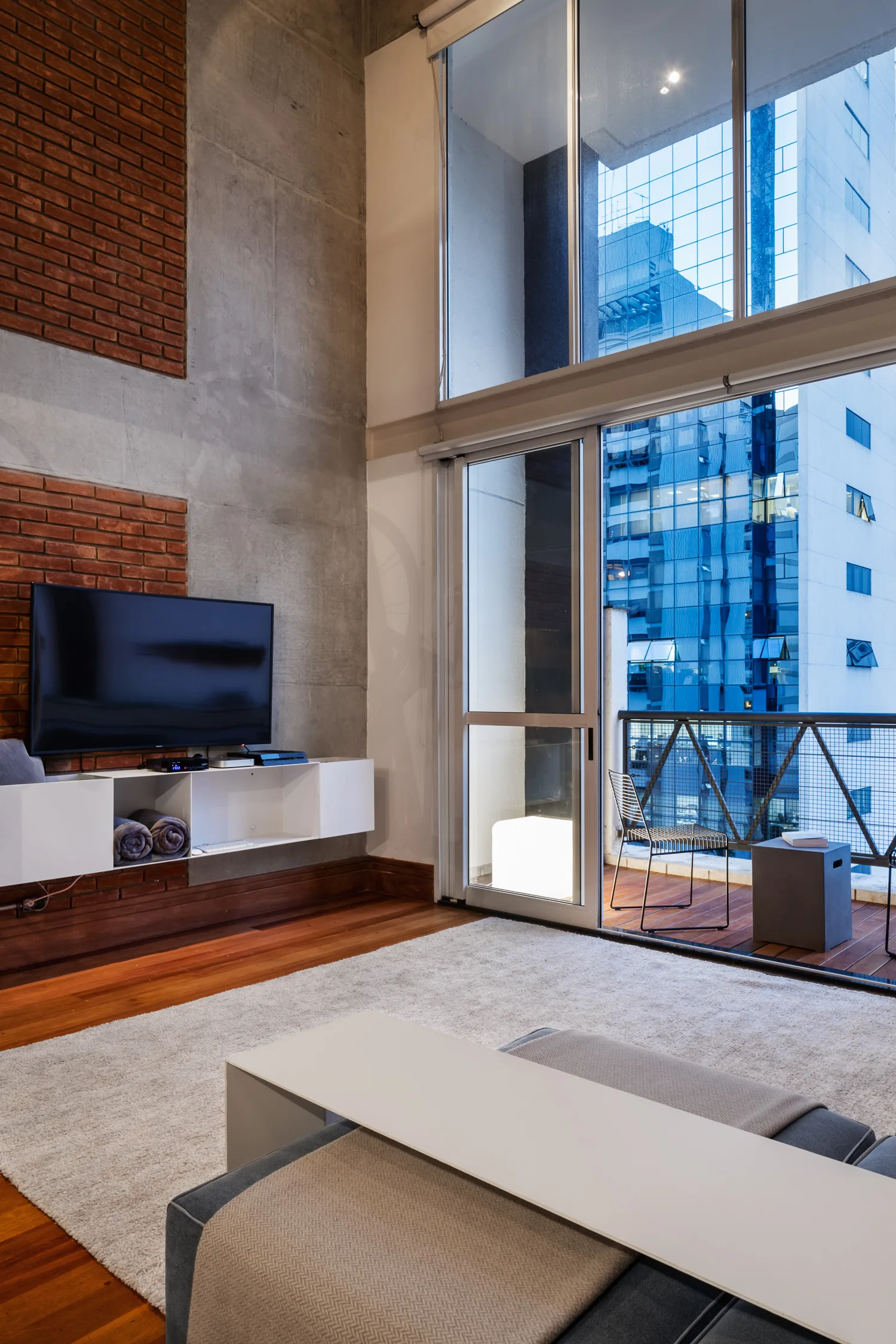 A modern living room features a white media console on a brick and concrete wall, alongside a large glass sliding door leading to a balcony.