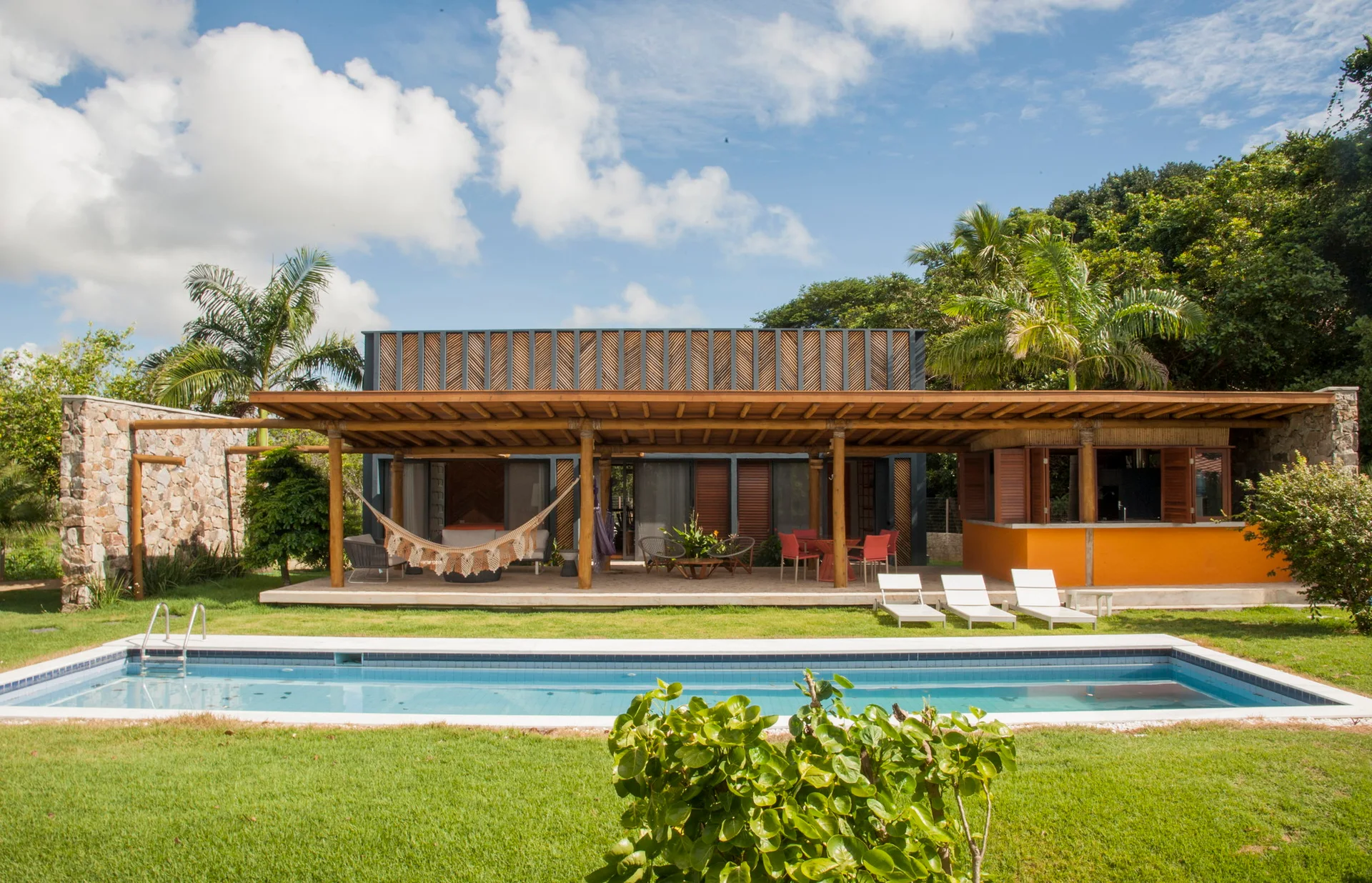 A rectangular swimming pool sits in front of a covered wooden patio featuring a hammock, dining set, orange bar, and stone wall.