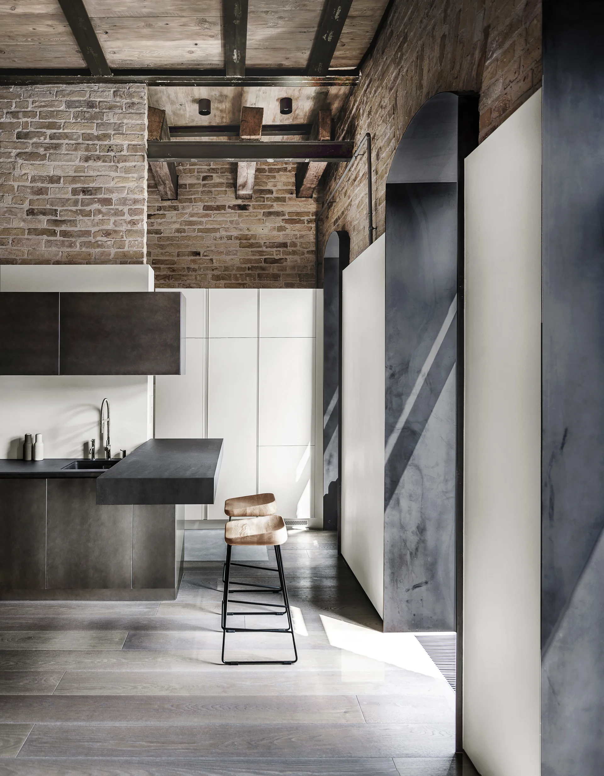A modern industrial kitchen features exposed brick walls, timber ceilings, a grey kitchen island with wood-topped bar stools, and dark metal archways.