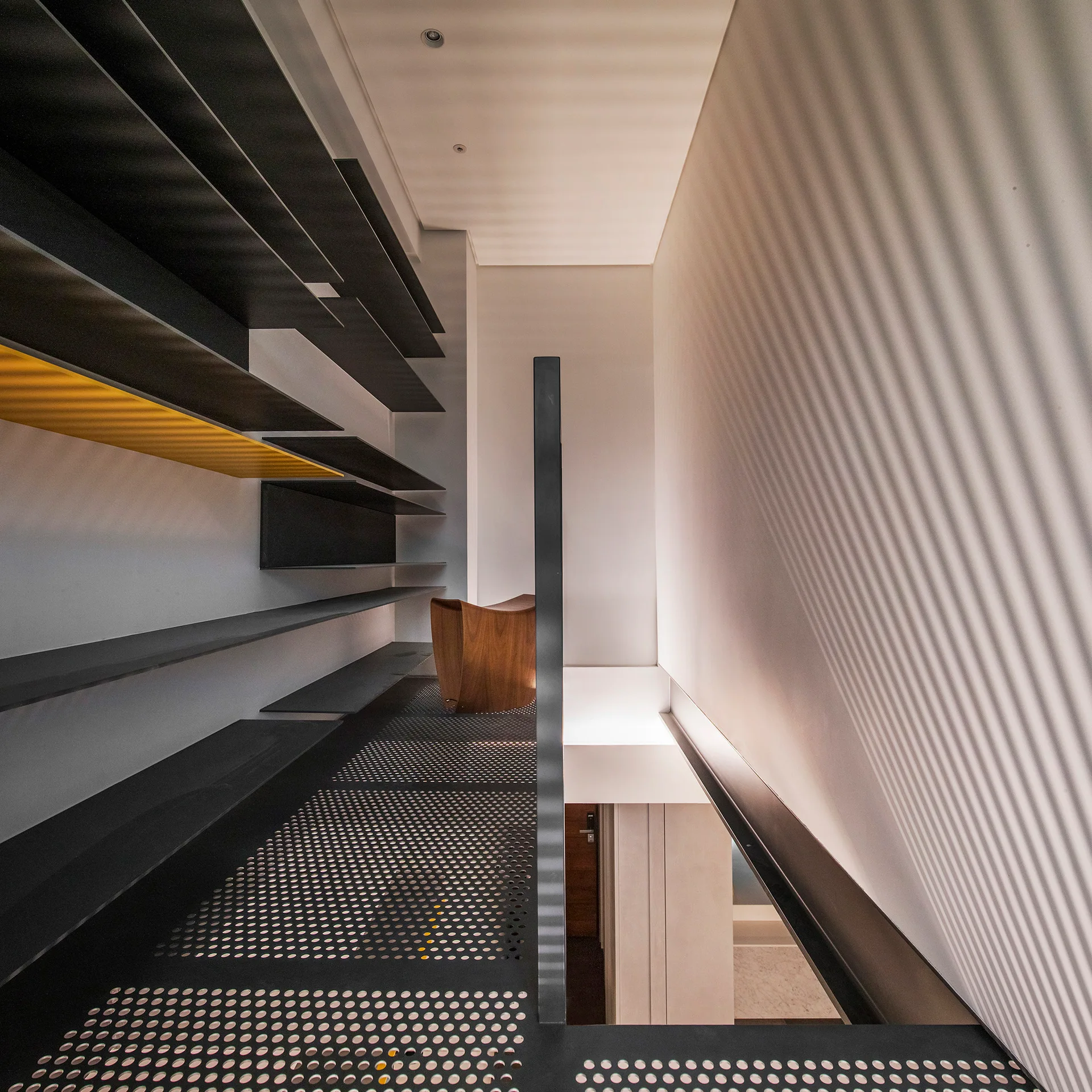 A minimalist mezzanine stair landing features a black perforated metal floor, dark wall-mounted shelving, and a single yellow accent shelf.