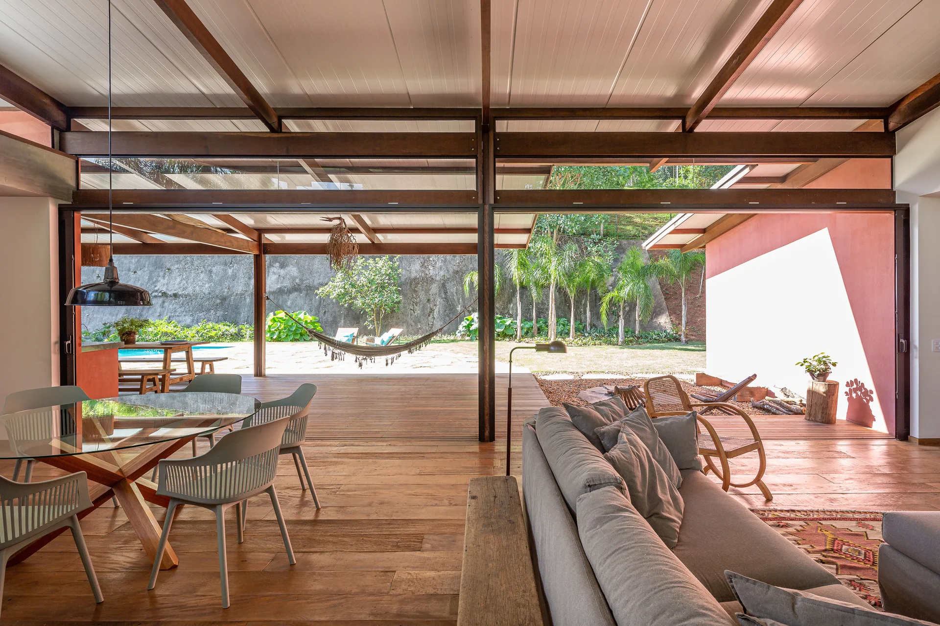 An open-concept living and dining space features exposed wooden beams, a glass-topped table, and large sliding doors opening to a garden patio with a hammock.