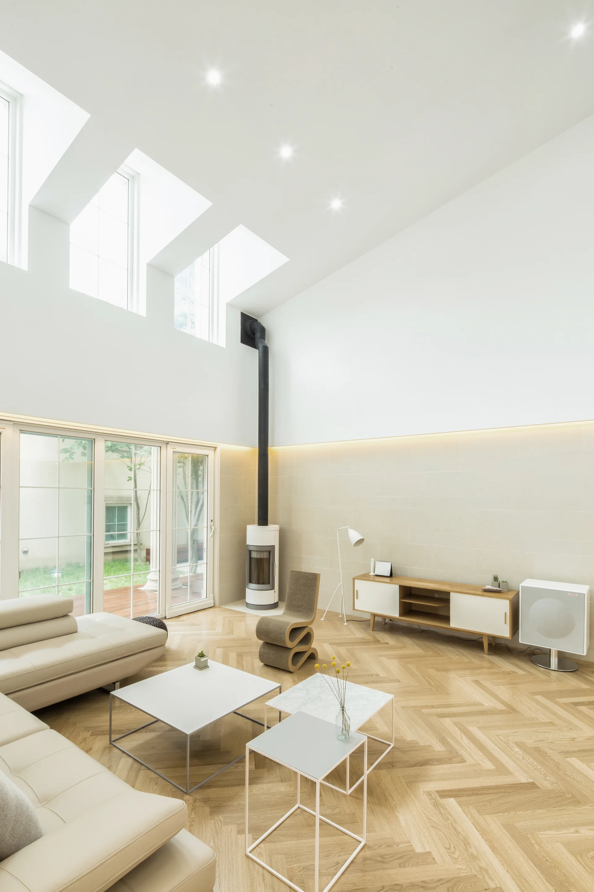 A bright living room features high ceilings with skylights, light wood herringbone flooring, a modular cream sofa, and a tall cylindrical fireplace.