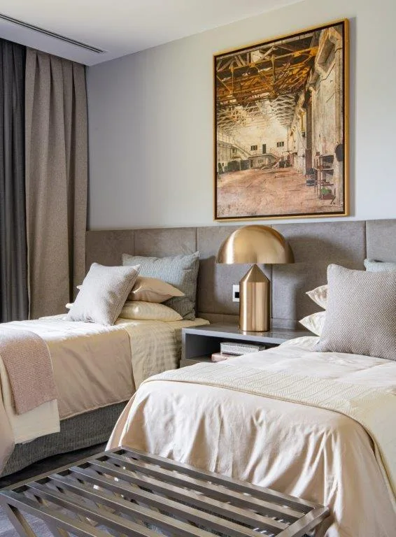 Two twin beds with beige linens sit on either side of a grey nightstand topped with a bronze dome lamp, backed by a large upholstered headboard.