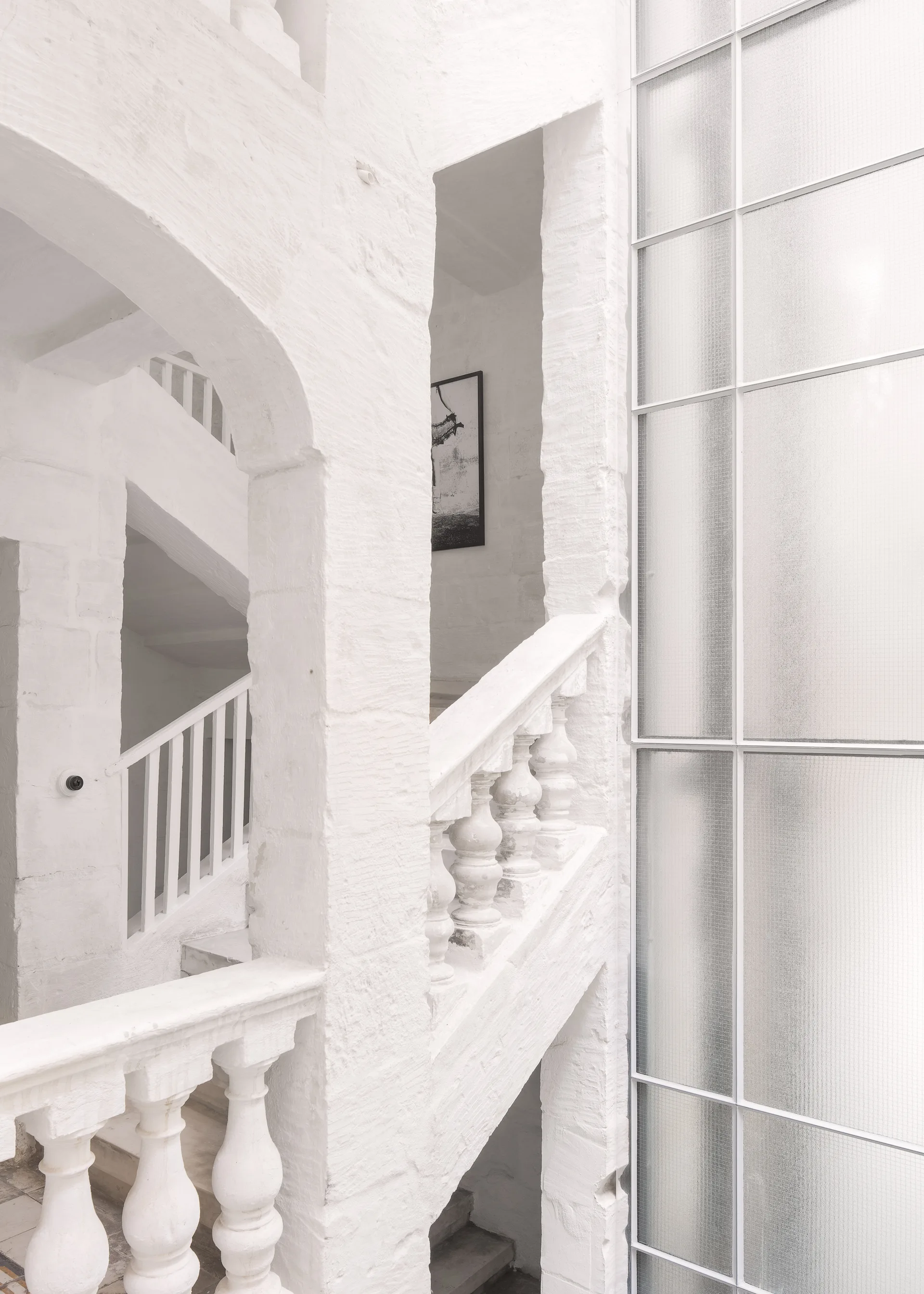 A multi-level white masonry staircase with classic stone balustrades features arched structural columns and a large frosted glass grid window.