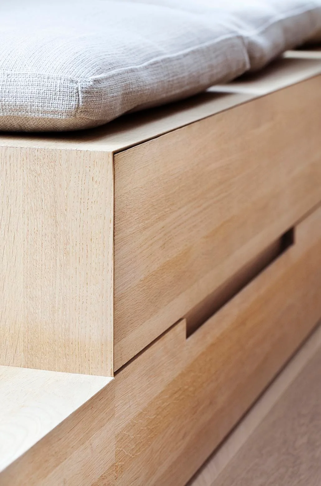 A close-up of a light wood storage bench featuring a textured cream cushion on top and a drawer with an integrated handleless slot.