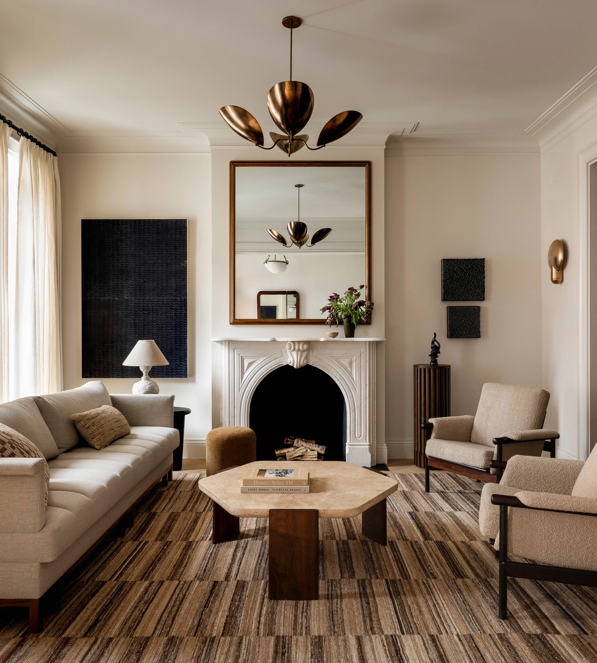 A neutral living room features a white stone fireplace, a hexagonal stone coffee table, two boucle armchairs, and a brass leaf chandelier.