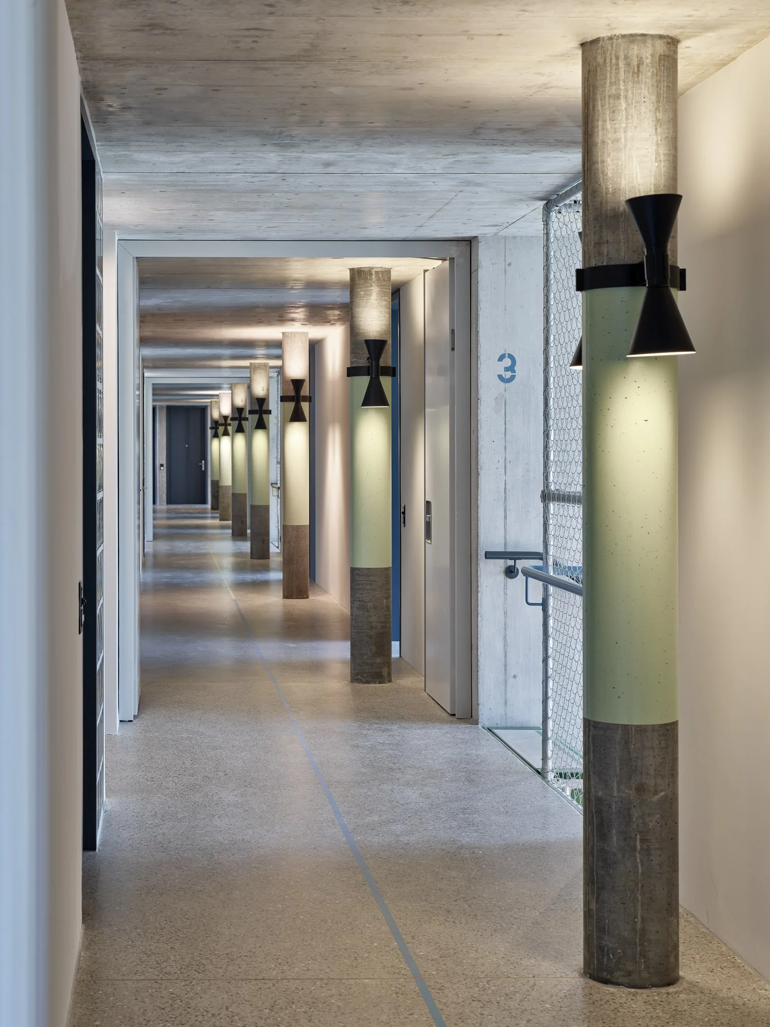 A long concrete hallway features a row of round columns painted green in the middle and mounted with hourglass-shaped black wall sconces.