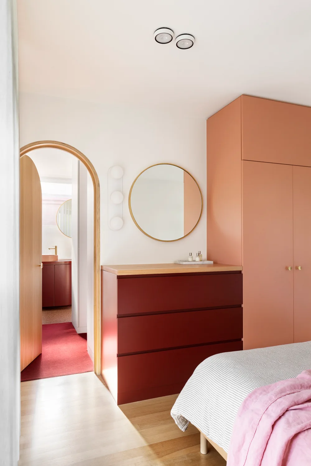 A contemporary bedroom features a peach-colored built-in wardrobe, a deep red dresser with a round gold mirror, and a wooden arched doorway.