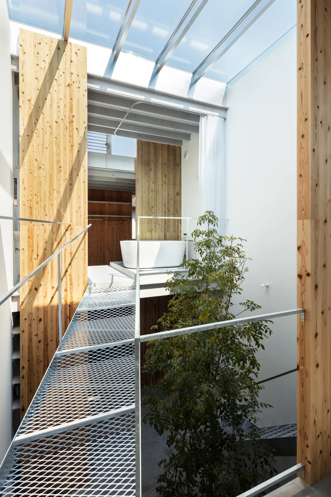 A modern minimalist bathroom featuring a white freestanding bathtub, an elevated metal grate bridge, and an indoor tree under a large glass skylight.