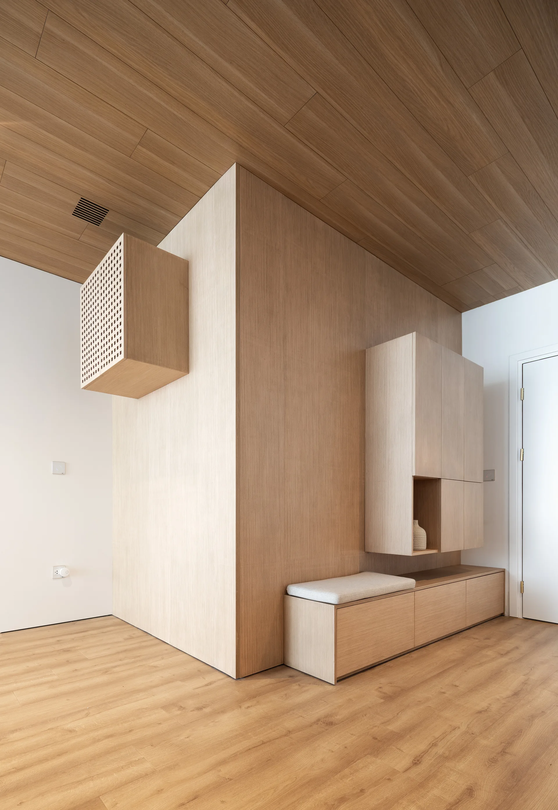 A minimalist entryway features custom light wood built-in cabinets, an integrated storage bench with a gray cushion, and a matching wood-planked ceiling.