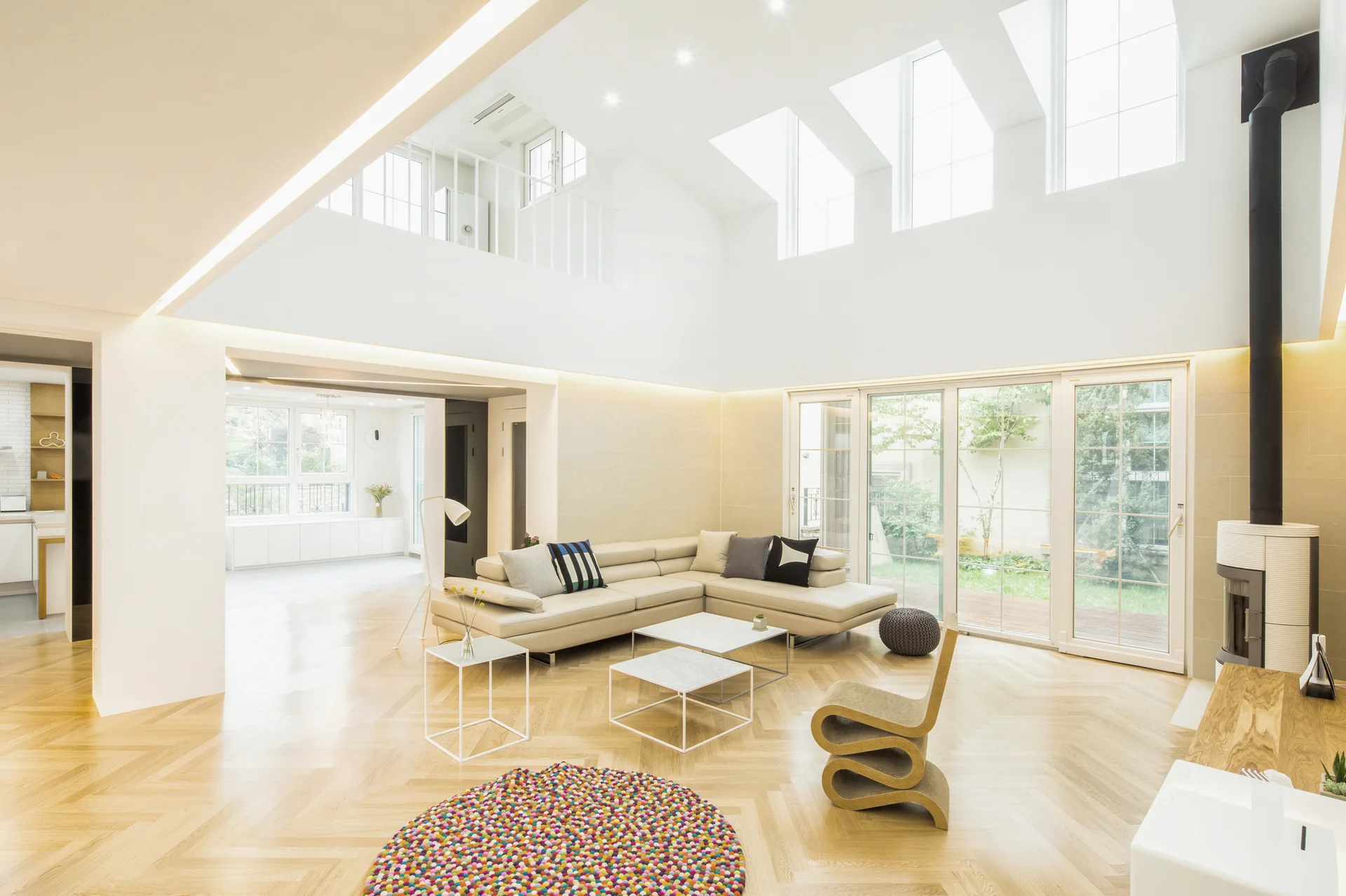 A modern double-height living room features a cream sectional sofa, herringbone wood floors, a sculptural chair, and a vaulted ceiling with skylights.