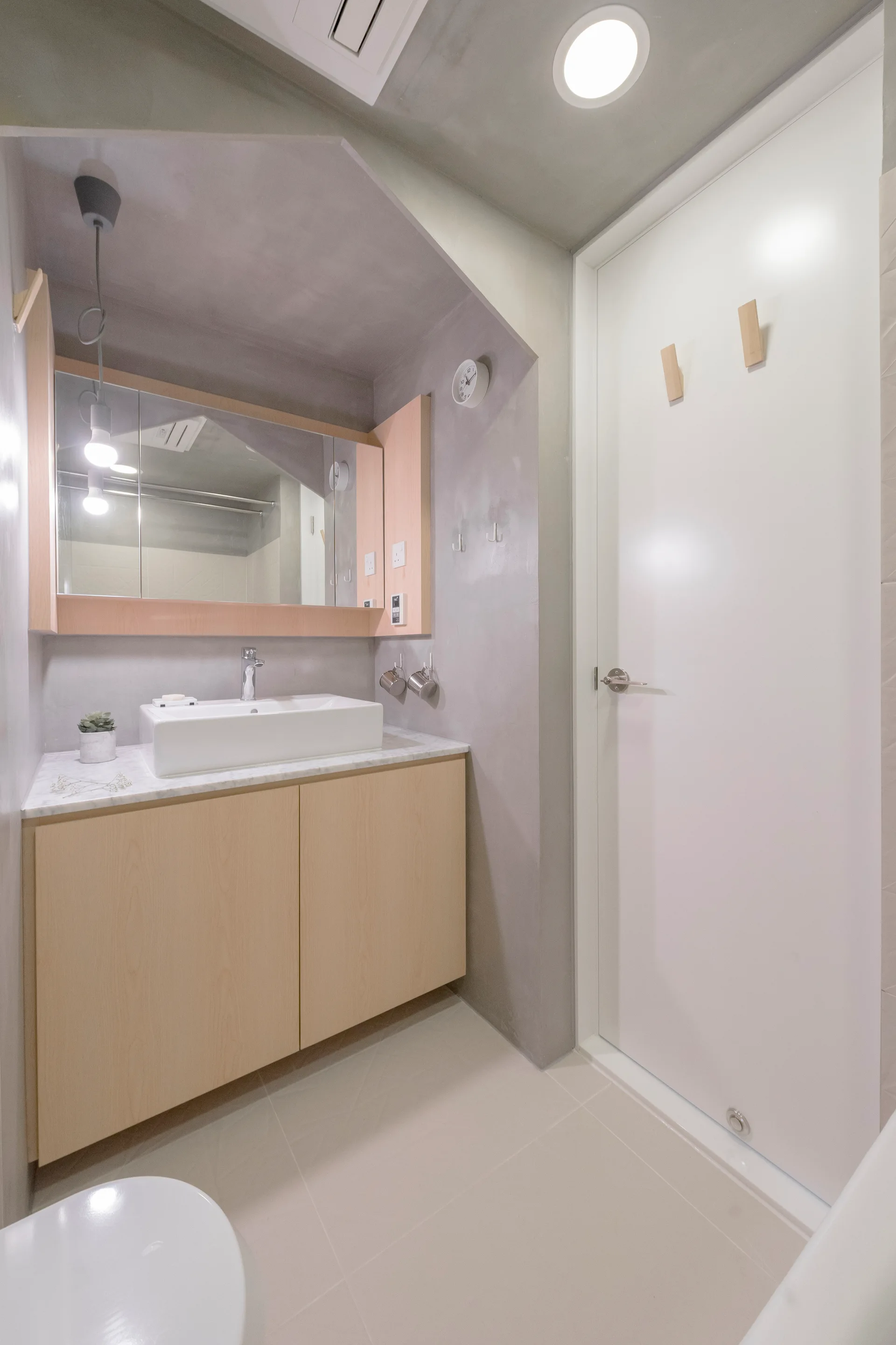 A minimalist bathroom showing a light wood vanity with a white sink, a mirror cabinet, grey plaster walls, and a white door.