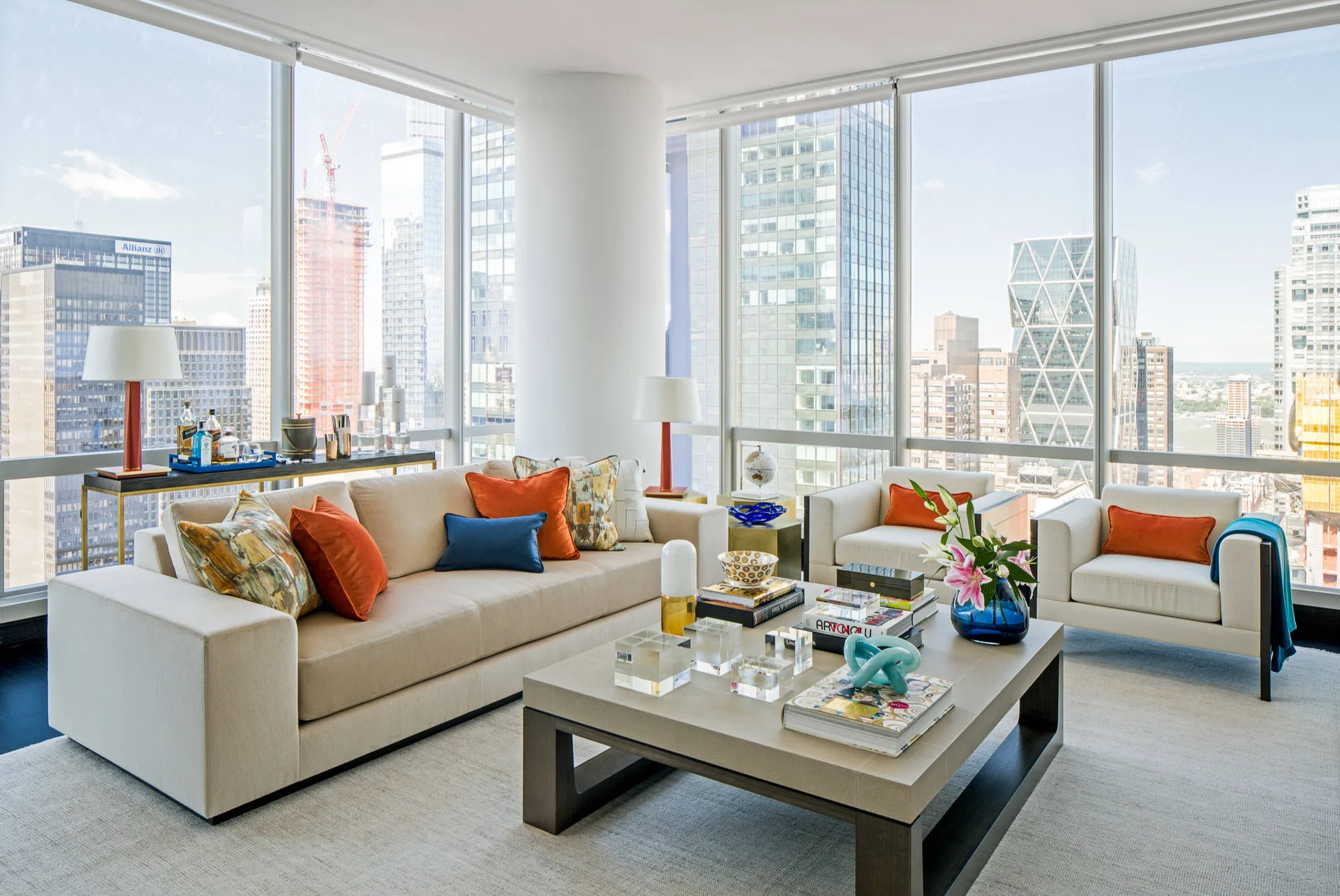 A modern high-rise living room with a beige sofa, two armchairs, a low coffee table, and large windows overlooking city skyscrapers.