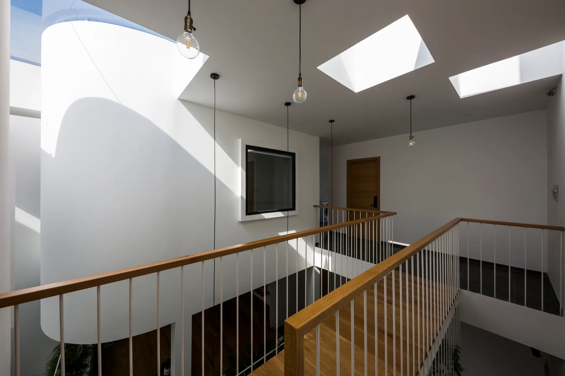 A bright upper hallway landing features a wooden handrail, thin white spindles, multiple ceiling skylights, and a prominent curved white wall.