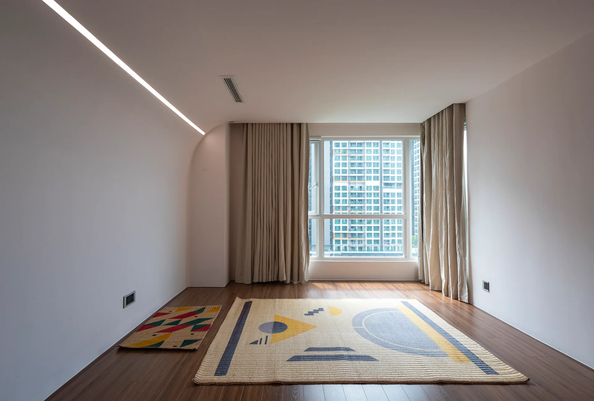 An empty room with dark wood floors, white walls, a linear light fixture, beige curtains, and a large geometric woven rug.
