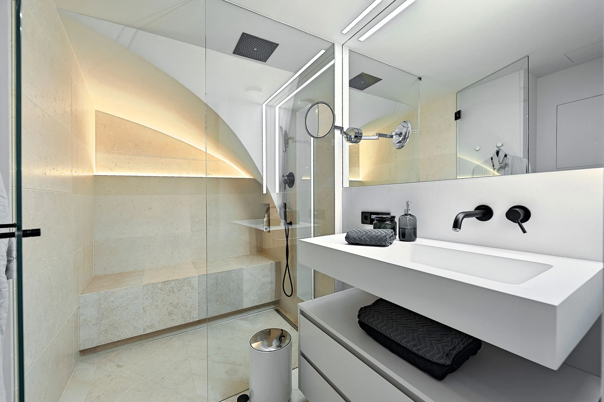 A modern bathroom features a glass-enclosed shower with beige stone tiles and a built-in bench, alongside a white floating vanity with black fixtures.