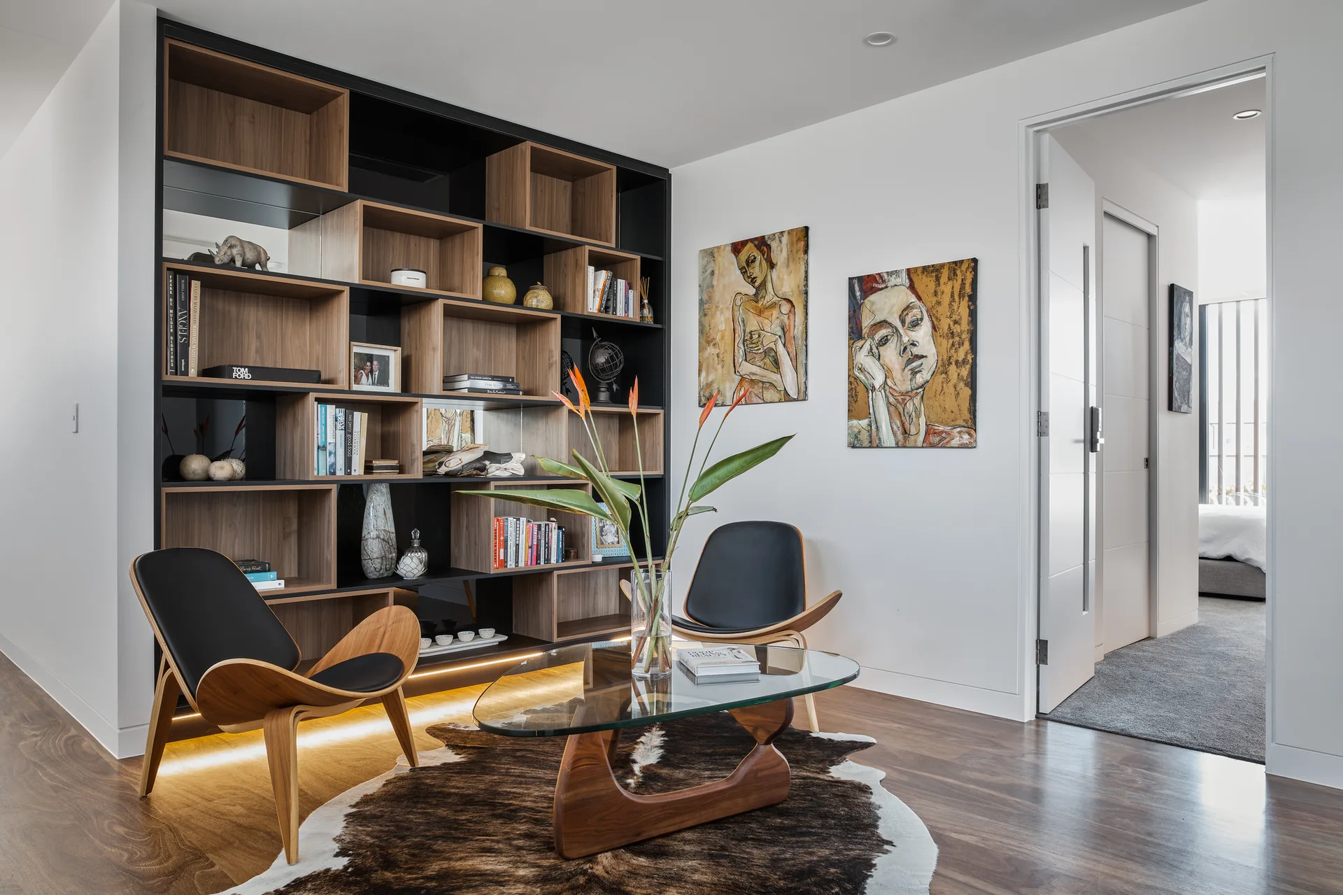 Two wooden shell chairs sit around a glass coffee table in a modern living room featuring a large built-in bookcase with warm accent lighting.