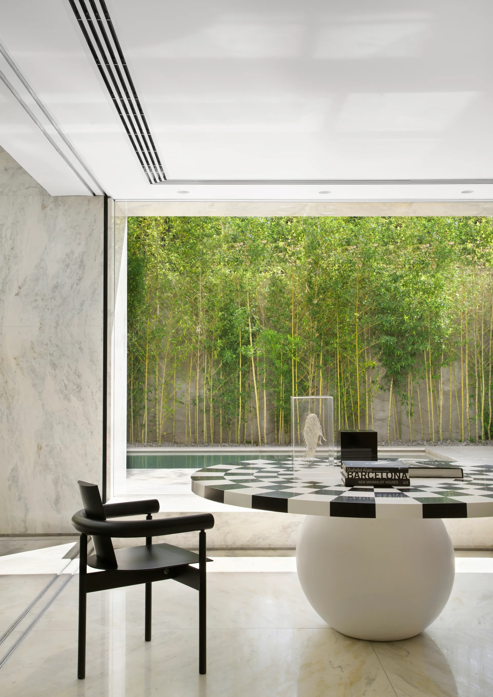 A modern room features a round black and white checkerboard table, a black armchair, and a glass wall looking out at green bamboo.
