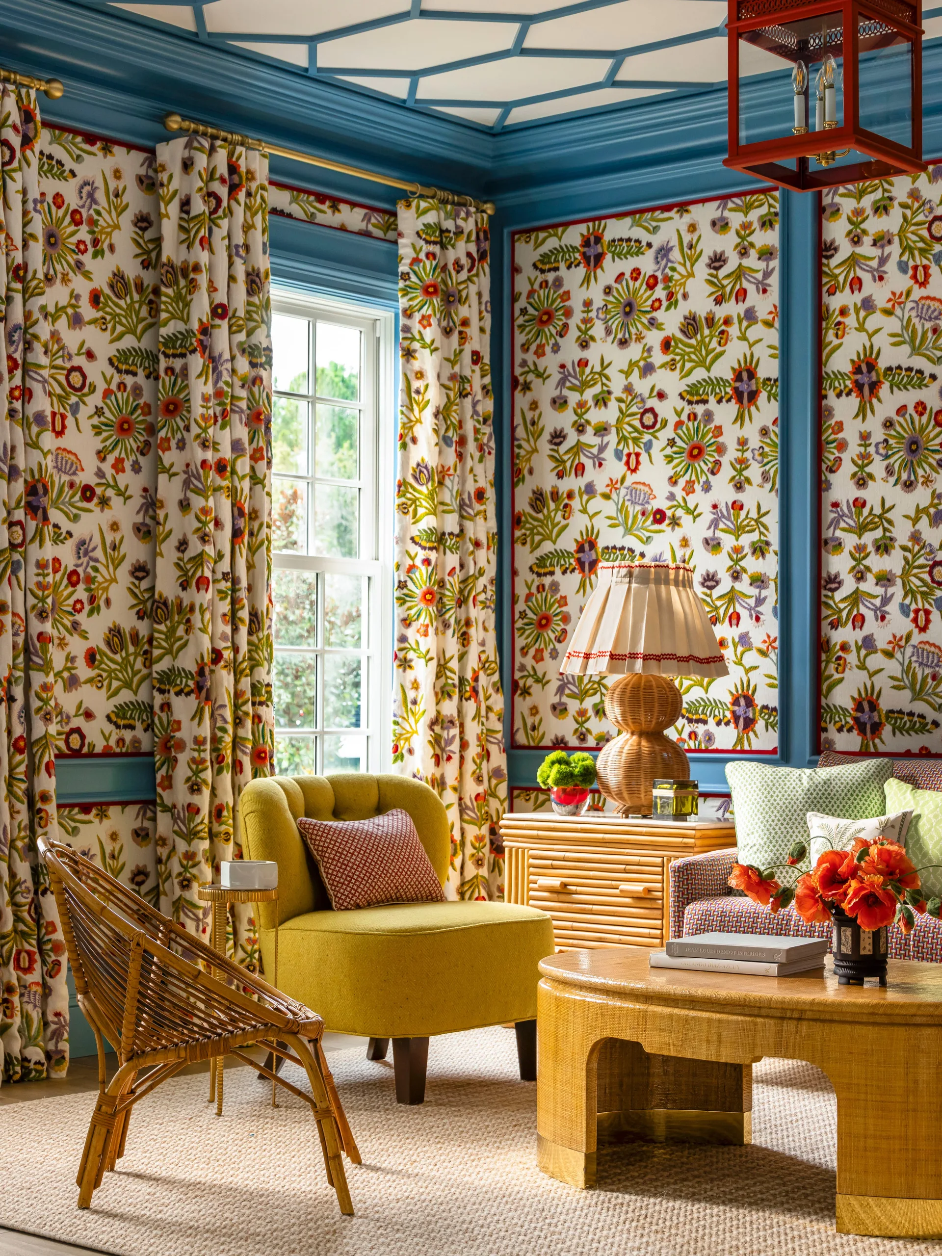 A maximalist living room featuring floral wallpaper framed by blue wood paneling, a yellow armchair, a rattan chair, and a geometric-patterned ceiling.